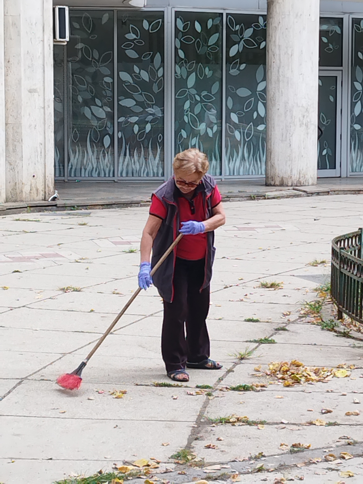 An elderly woman with blonde hair and glasses is sweeping a paved area with a red-bristled broom. She is wearing a red shirt, a dark vest, dark pants, and blue gloves. The pavement is made of rectangular concrete slabs and is littered with fallen yellow and brown leaves. In the background, there is a building with large glass windows decorated with a repeating pattern of leaves and grass. Two large concrete pillars stand in front of the building. To the right, a section of a green metal fence is visible. The scene appears to be an outdoor public space. The presence of fallen leaves suggests it is autumn. There is no discernible indication of this being in Bucharest, Romania.