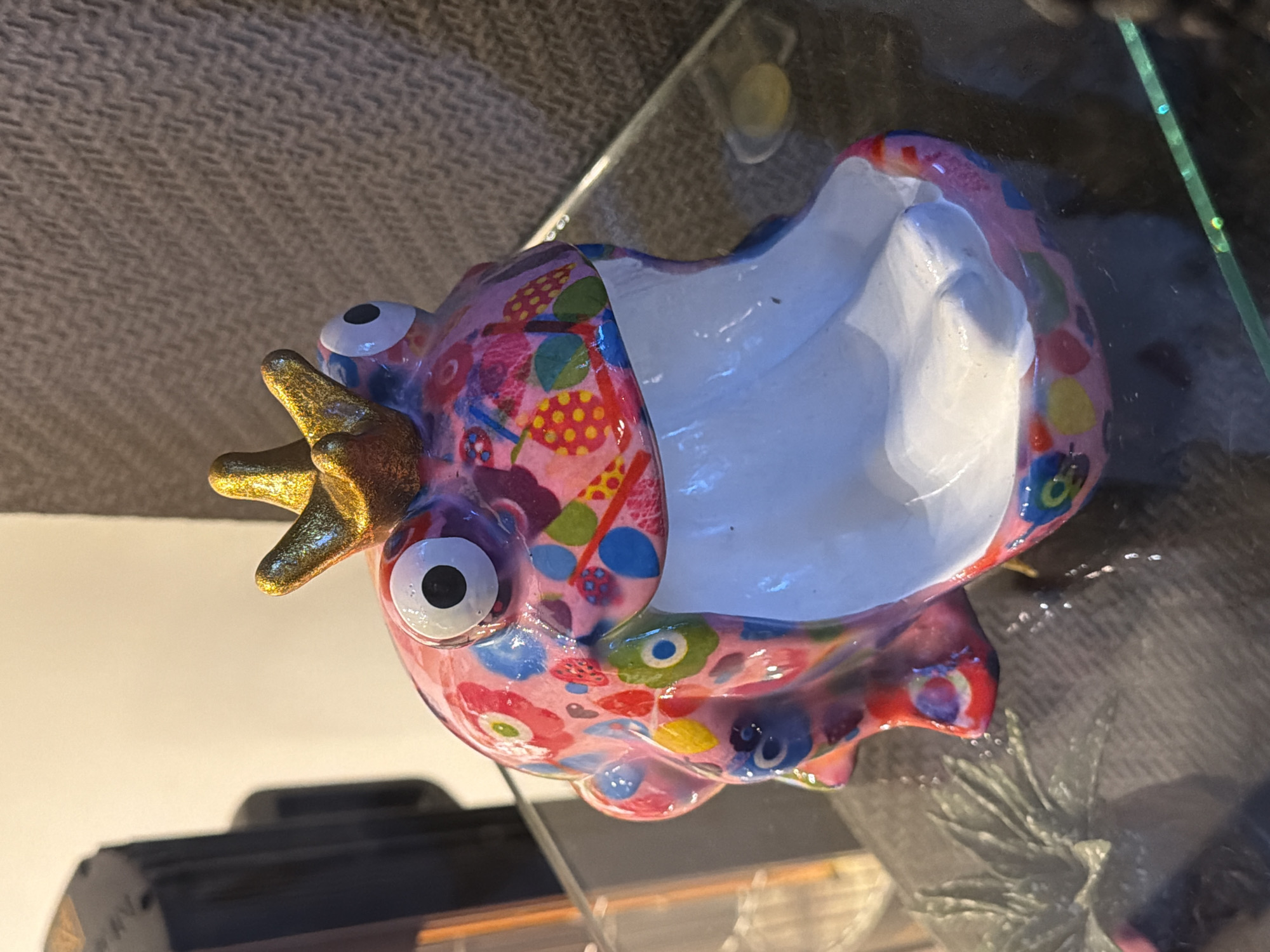 The image displays a close-up, eye-level shot of a pink ceramic figurine shaped like a creature with a wide, open mouth and a golden crown. The figurine is decorated with colorful floral and abstract patterns in shades of blue, red, yellow, green, and white. It has two large, googly eyes with black pupils. The figurine is positioned on a clear glass surface, which reflects light and some of the surrounding environment. Behind the figurine, a textured, dark brown wall or fabric is visible. To the left, a dark, rectangular object with some reflective elements is partially in frame. The overall lighting is soft, with a warm tone. The image's composition focuses primarily on the figurine. The location in Üsküdar, Turkey, is not discernible from the visual information provided in the image.