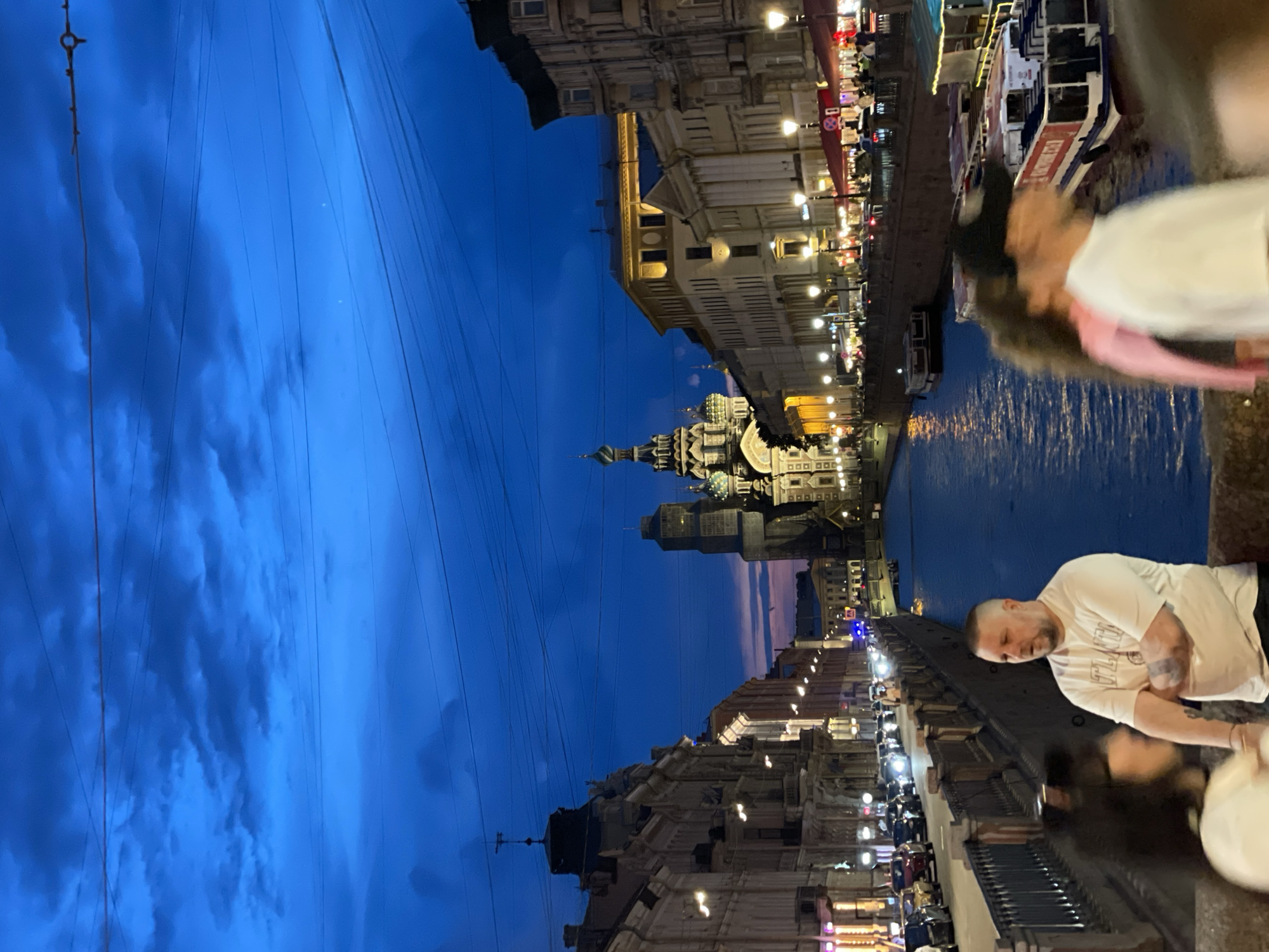 This image captures an urban scene in Saint Petersburg, Russia, during twilight or early night, looking down the Griboyedov Canal towards the Church of the Savior on Spilled Blood. The sky is a deep, rich blue with scattered dark clouds, transitioning to a lighter purplish hue near the horizon on the left. Numerous dark overhead utility wires crisscross the upper portion of the frame.
The central feature in the mid-distance is the illuminated Church of the Savior on Spilled Blood, with its distinctive onion domes and ornate architectural details. Flanking the canal on both sides are multi-story buildings of varying architectural styles, predominantly stone or stucco, illuminated by numerous streetlights and internal lights, creating warm glows. On the left bank, a continuous line of vehicles, some with headlights visible, suggests active traffic. People are visible along the sidewalks, dining in outdoor seating areas, and walking.
The Griboyedov Canal occupies the center of the lower frame, its surface reflecting the ambient lights and appearing dark blue. Several boats, including at least two white and red tour boats, are docked along the right bank of the canal.
In the immediate foreground, a stone bridge or embankment serves as the vantage point. Three individuals are prominently featured. On the left, a person with dark hair is partially visible and out of focus, facing right. To their right, a bald man with a short beard, wearing a light-colored t-shirt (possibly bearing "ATLN" text) and dark pants, stands with his arms crossed, looking slightly left. A tattoo is visible on his left forearm. On the far right, two blurry figures are in motion; one wears a pink top, and the other wears a white t-shirt and a dark baseball cap. The scene is brightly lit by artificial sources, contrasting with the deep blue of the sky and canal.