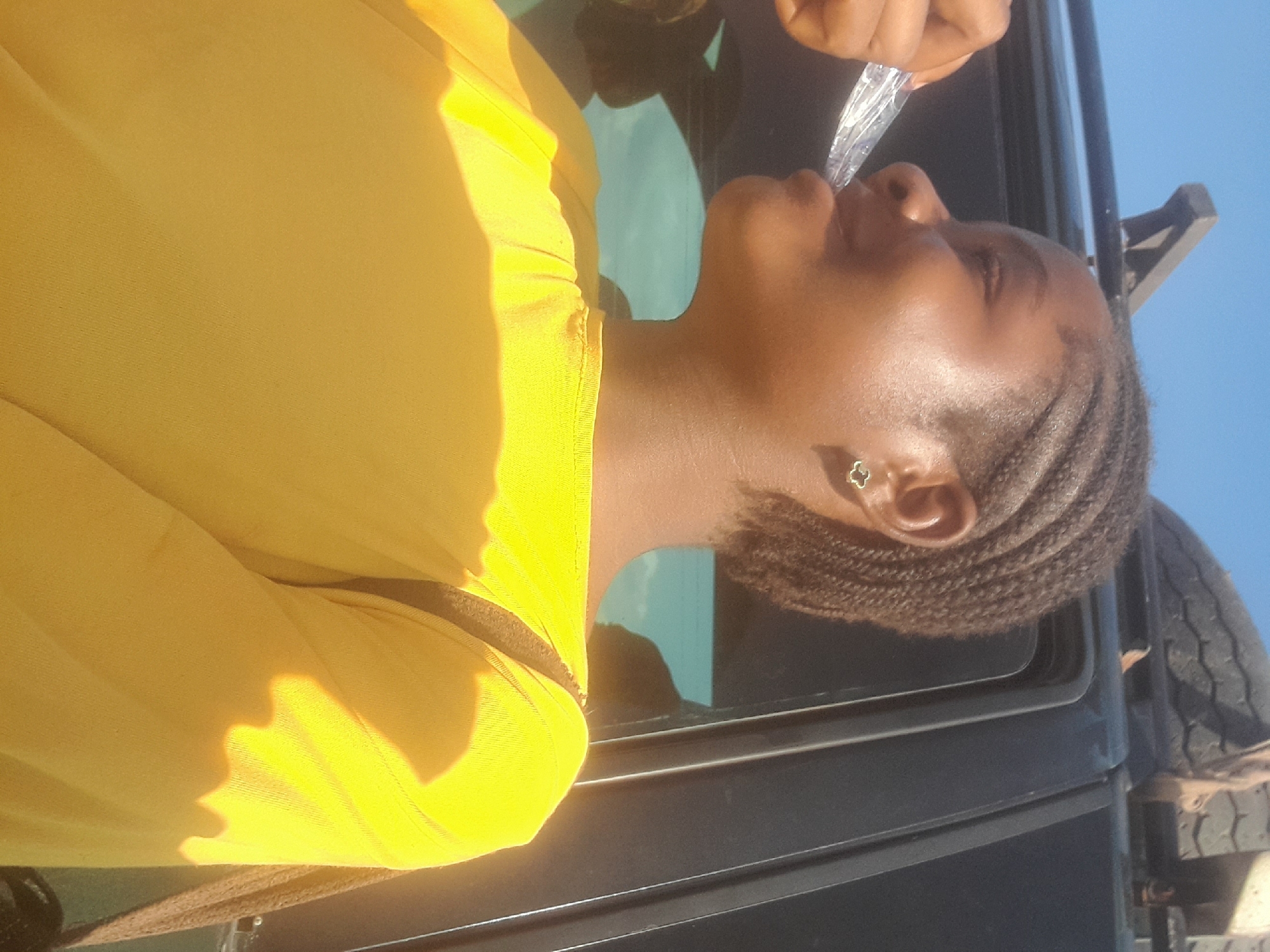 In Ibi, Nigeria, a young woman with braided hair is photographed from the chest up, angled to her right. She is wearing a bright yellow long-sleeved shirt. Her right hand is near her face, holding a clear plastic object, possibly a bag of chips or a snack. She appears to be eating or about to eat from it. A small, gold-colored earring shaped like a clover or flower is visible in her left ear. The background shows the dark exterior of a vehicle, with a spare tire and roof rack visible on top. The sky is a clear blue. The lighting suggests it is daytime, with sunlight casting shadows on the woman's shirt.