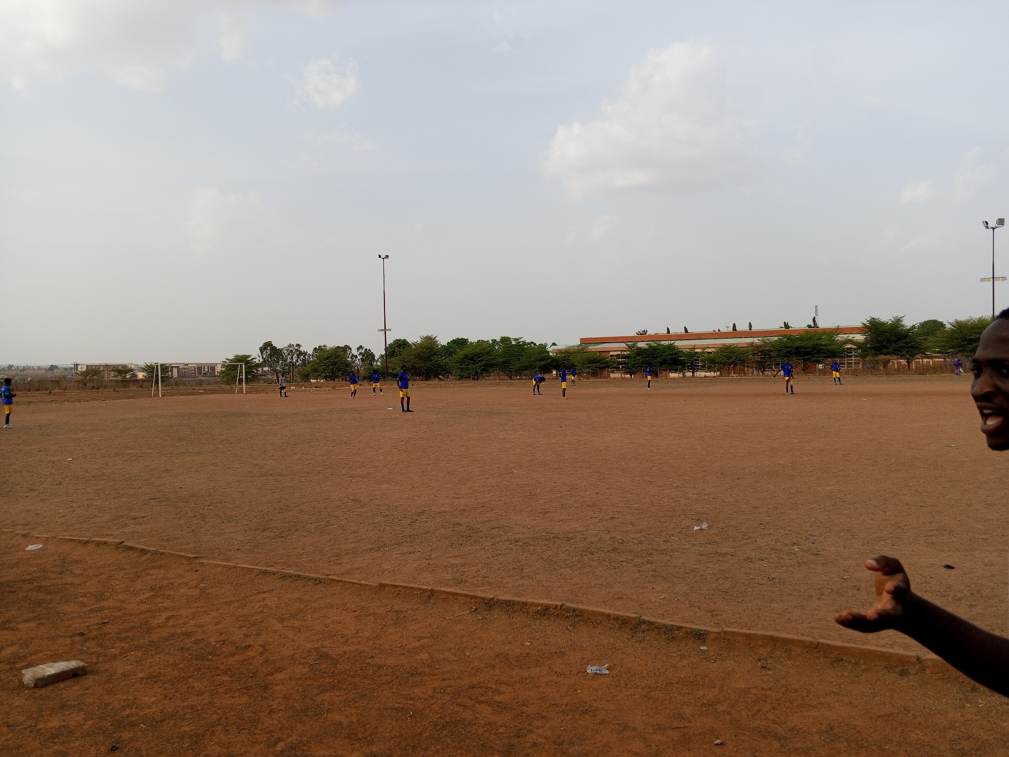 The image shows a dirt soccer field in Minna, Nigeria. The field is dry and barren, with scattered debris. A group of soccer players wearing blue and yellow uniforms is practicing on the field. In the background are trees and a low-rise building. A person is partially visible in the right foreground, gesturing with their hand. The sky is overcast. Two light poles are visible in the mid-ground.