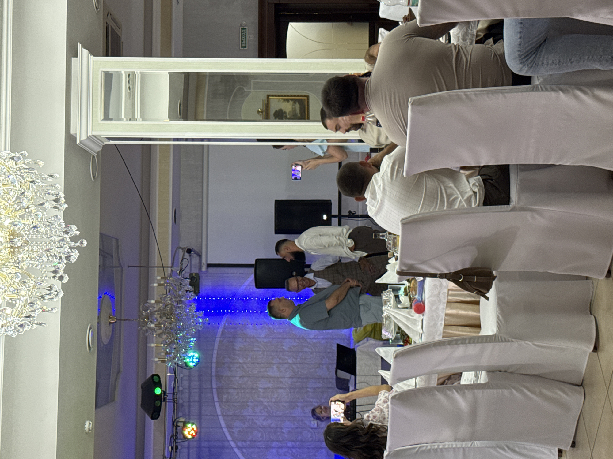 The image depicts an indoor event, likely a banquet or reception, in Ust-Labinsk, Russia. The room is adorned with ornate crystal chandeliers and decorative lighting, suggesting a formal setting. Several people are present, seated at tables covered with white tablecloths and dressed in formal or semi-formal attire. In the background, three men are engaged in conversation, one of whom is holding a phone, possibly taking a picture or video. Another woman in the foreground is also holding a phone and appears to be recording the scene. The overall atmosphere appears festive and social, with guests interacting and documenting the occasion. The presence of a "ВЫХОД" (Exit) sign indicates a public venue.
