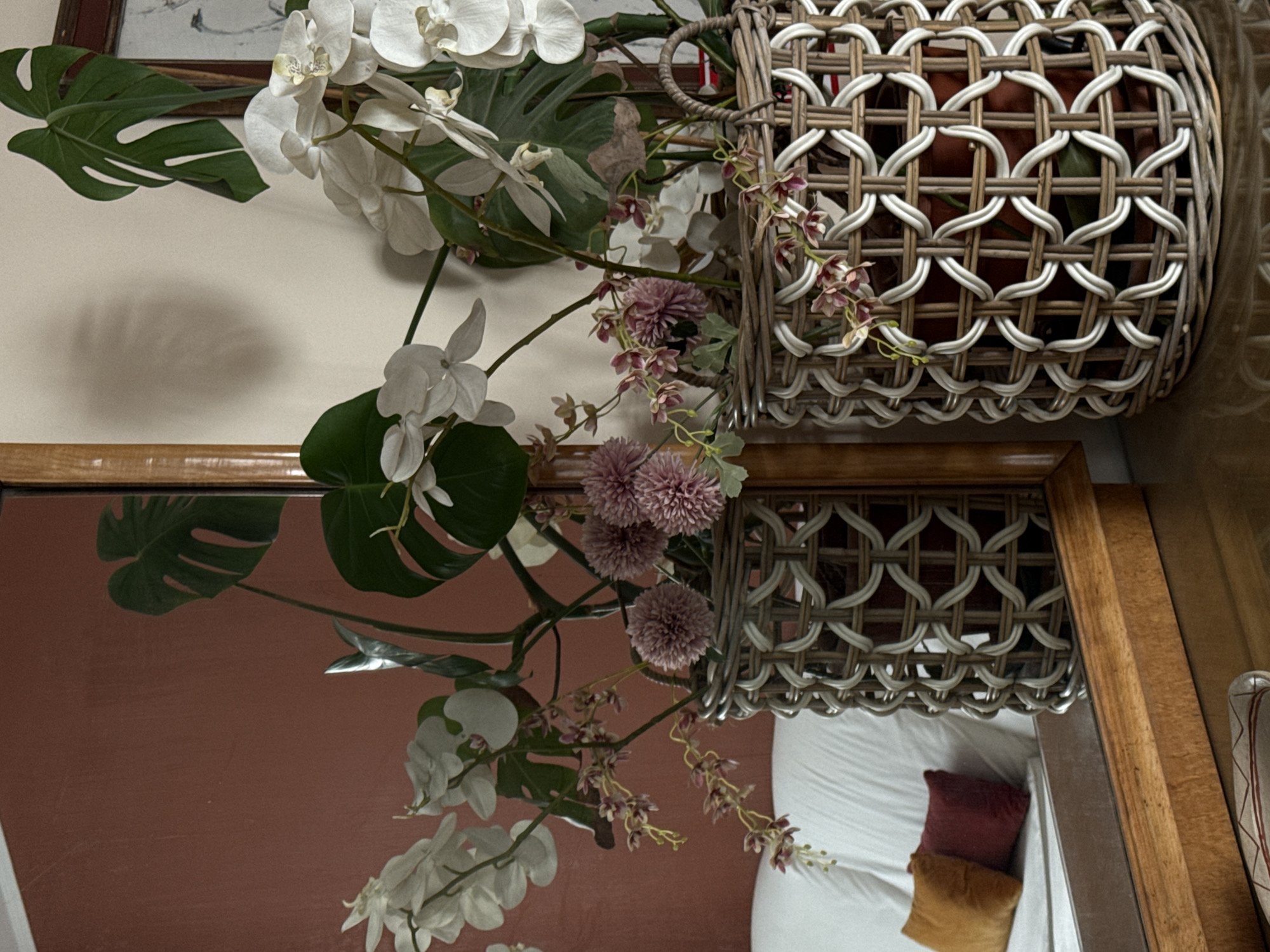 The image depicts a still life arrangement indoors, consisting of a woven basket filled with various floral and foliage elements. Prominent are white orchids and Monstera leaves. The entire arrangement is positioned above a framed mirror, reflecting the scene below, which includes a bed with white linens and pillows.
Without additional information, I cannot determine the precise location of the image in Milan, Italy.