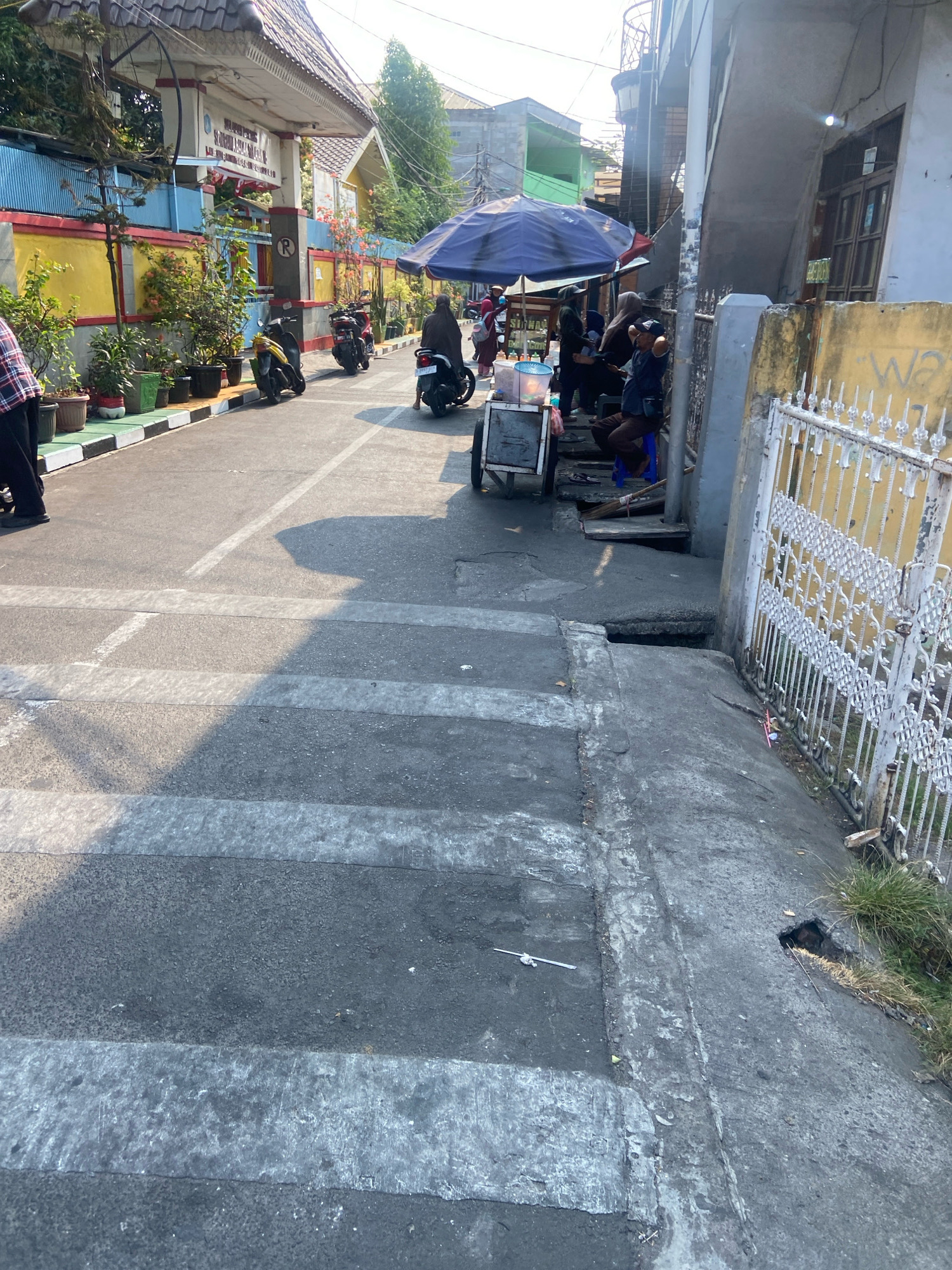 The image depicts a street scene in North Jakarta, Indonesia, under a bright, sunny sky. The foreground features a weathered asphalt road with distinct white pedestrian crossing markings. To the left, a row of potted plants lines a sidewalk in front of a building with a yellow and red facade. Behind the plants, a blue wall is visible, and further back, the entrance to a building is adorned with a grey tiled roof and decorative trim. Several motorcycles are parked on the side of the road.
In the middle ground, an open-air market is set up. A large blue umbrella provides shade for several people and a food cart. A few individuals are gathered around the cart, and one person is seated on a blue stool. A person on a motorcycle is seen moving away from the market area. The street continues to the right, leading to a grey building with a yellow wall and a white ornate metal gate. The ground on the right side of the street shows signs of disrepair, with a hole in the pavement near the gate. The overall impression is a busy, functional street with elements of commerce and everyday life.