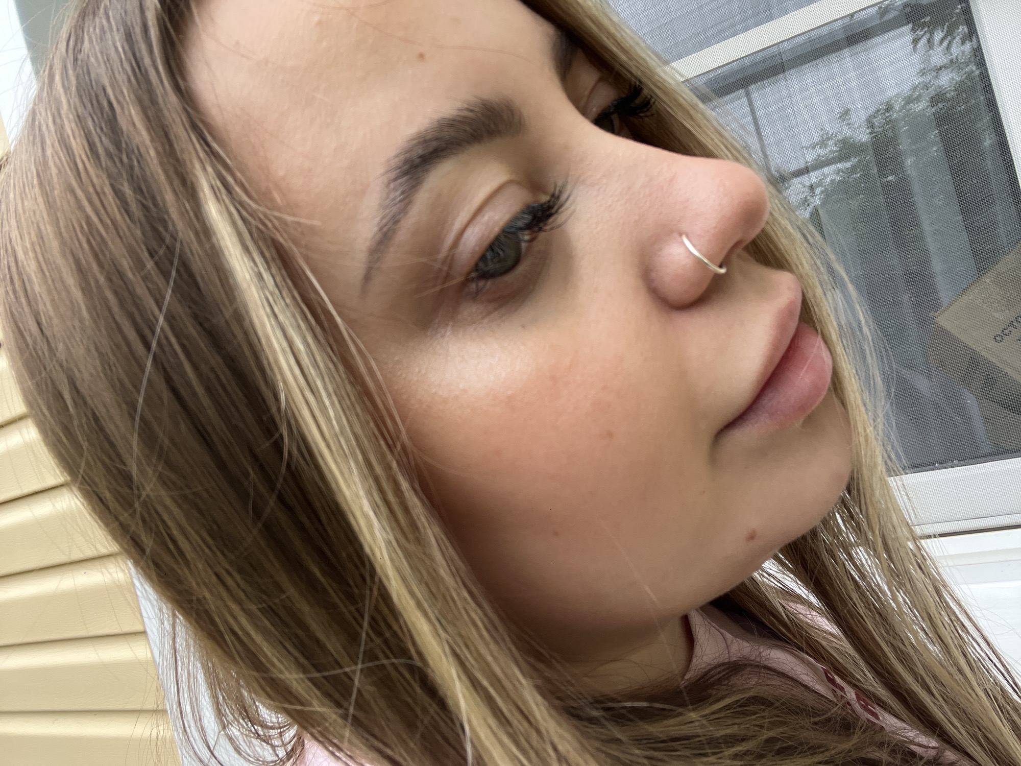 A close-up, angled portrait captures a woman with light brown hair with blonde highlights. She has a silver nose ring and dark eyelashes. Her skin tone appears fair, and she is wearing a light pink top. Behind her, a window screen is visible, as well as a light yellow exterior wall.
