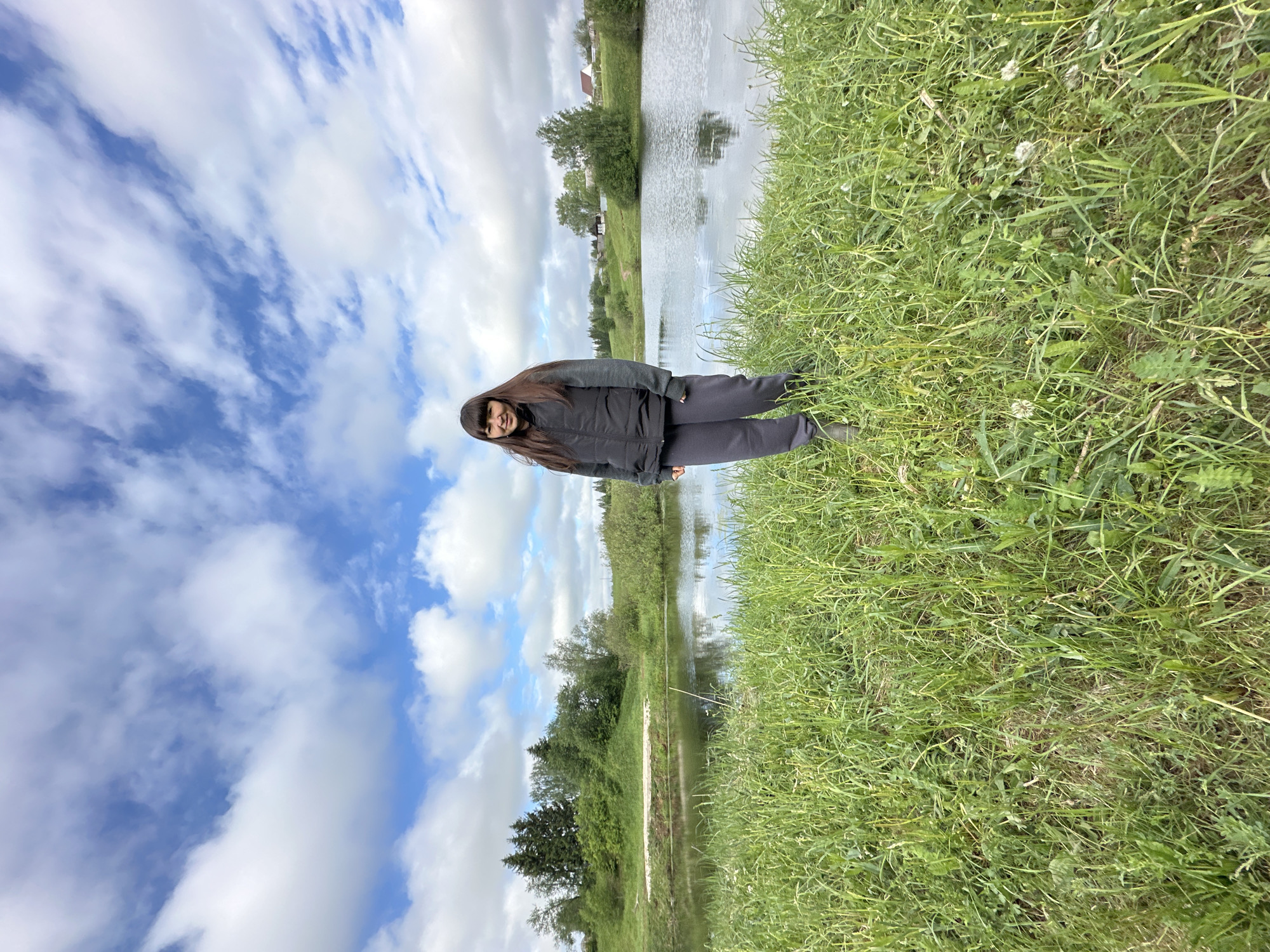 The image depicts a young woman standing near a body of water, likely a pond or a narrow river, in Среднее Кечево, Russia. She is wearing a dark jacket and pants, and her dark hair is long. The background shows a green landscape with trees and grass, with the body of water reflecting the sky. The sky is partly cloudy. The photograph appears to have been taken at a tilted angle. The woman appears to be standing on a grassy bank.