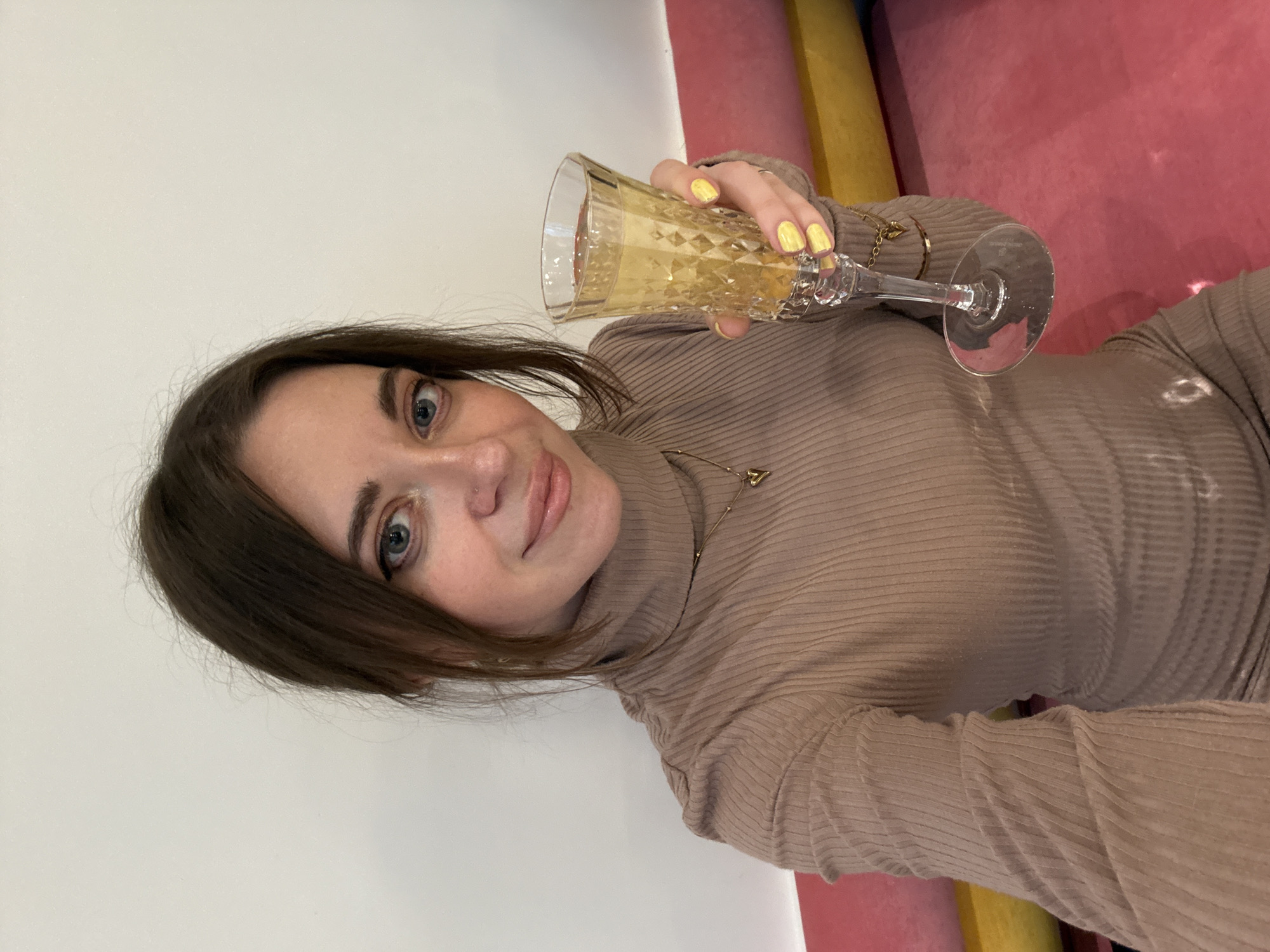 A woman is holding a glass of a yellow liquid. She is wearing a beige ribbed turtleneck sweater and a delicate gold necklace. Her nails are painted bright yellow. The background shows a section of a wall painted white, a vertical section colored yellow, and a pink wall. The city is Moscow, Russia.