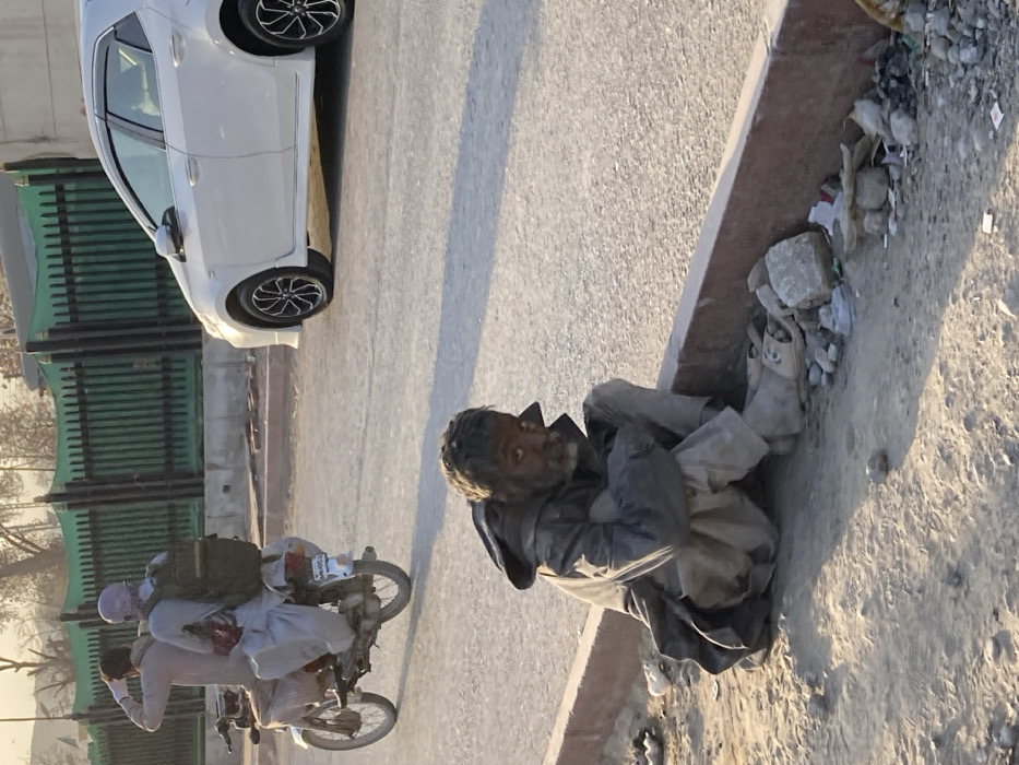 In Quetta City Tehsil, Pakistan, an unkempt, elderly male subject with a beard and disheveled dark hair is seated on a dirt shoulder beside an asphalt road, looking directly forward. He wears a dark, soiled jacket over light-colored trousers and open-toed footwear. His knees are drawn up, and his arms are clasped. Adjacent to him is a red-topped concrete curb, with scattered debris and small rocks on the dirt. On the asphalt road, a white Toyota Corolla sedan with black and silver multi-spoke rims is partially visible on the right. To the left, a silver motorcycle carries two male subjects. The passenger, wearing a light head covering and a dark backpack, is looking back over his right shoulder. The driver and passenger are dressed in light-colored traditional attire. In the background, a green metal fence runs parallel to the road, with sparse trees visible beyond. The scene is illuminated by natural light, casting subtle shadows, suggesting late afternoon or early morning conditions.