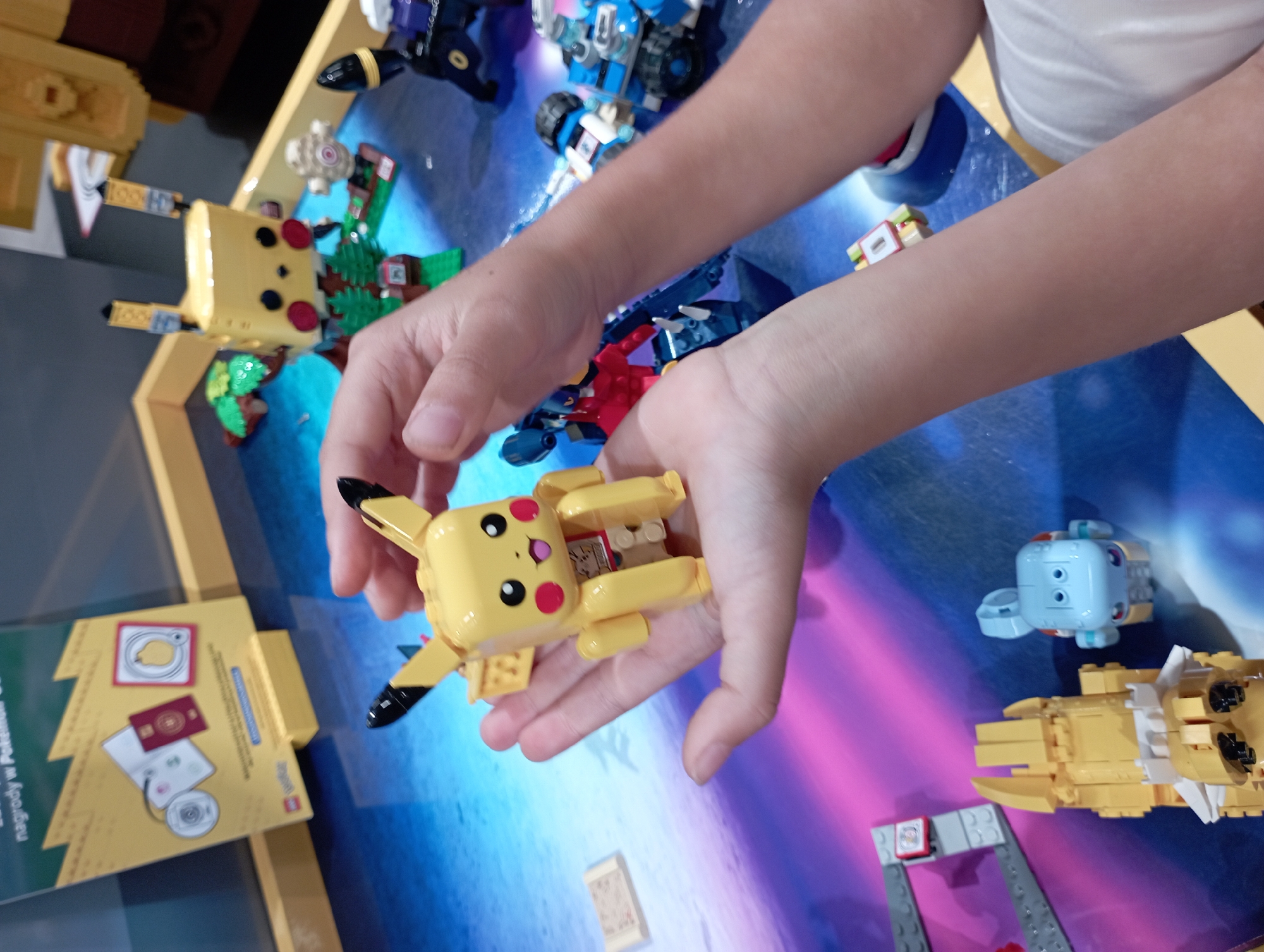 The image depicts a close-up view of hands holding a yellow LEGO Pikachu figure. The Pikachu figure is detailed, with black-tipped ears, red cheeks, and a smiling expression. It is positioned on the palm of a person's left hand, while their right hand is visible to the right, supporting the Pikachu from behind.
In the background, several other LEGO figures and structures are visible. These include a larger, yellow LEGO Pikachu-like character with green foliage, a blue LEGO robot-like figure, and other smaller LEGO elements. The surface beneath the LEGOs appears to be a blue table with a colorful, abstract design that transitions from pink to purple. The overall setting suggests an indoor environment, likely a display or play area.
This scene is consistent with the LEGO Store at the Silesia City Center mall in Katowice, Poland. The mall, located at Chorzowska 107, 109-111, 40-101 Katowice, Poland, is a major retail and entertainment complex that houses a LEGO Store, known for its interactive displays and themed exhibits, including those featuring popular characters like Pokémon. The presence of LEGO merchandise and the vibrant, child-friendly atmosphere strongly indicate this location.