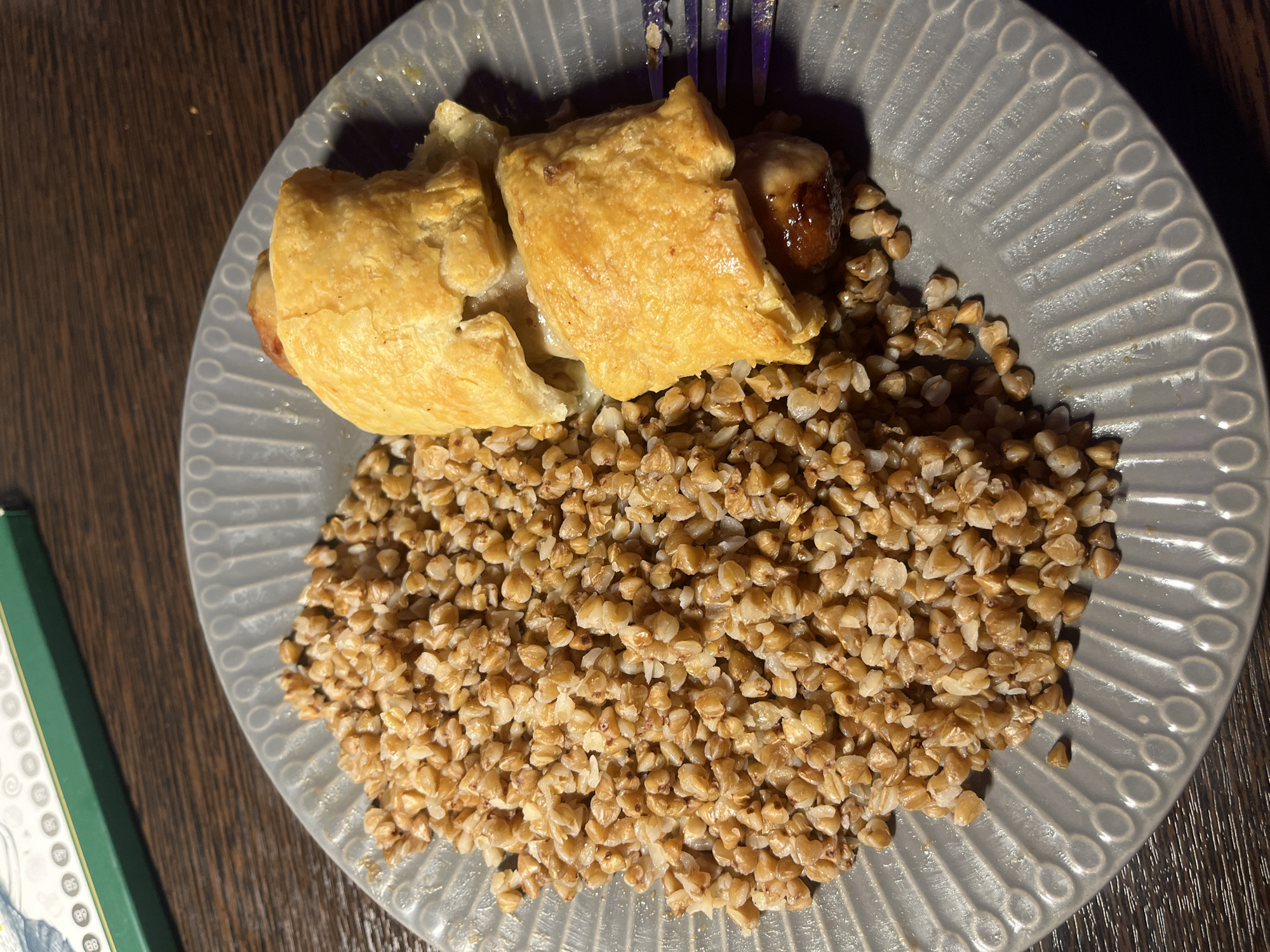The image shows a plate of food on a wooden surface. The plate, a grey, decorative one, contains a serving of buckwheat and two pastries. The pastries appear to be filled with meat, possibly sausage. A set of cutlery is slightly visible next to the pastries. The scene appears to be indoors, possibly in a private residence. It is not possible to determine the location in the city of Saint Petersburg, Russia, from this image.