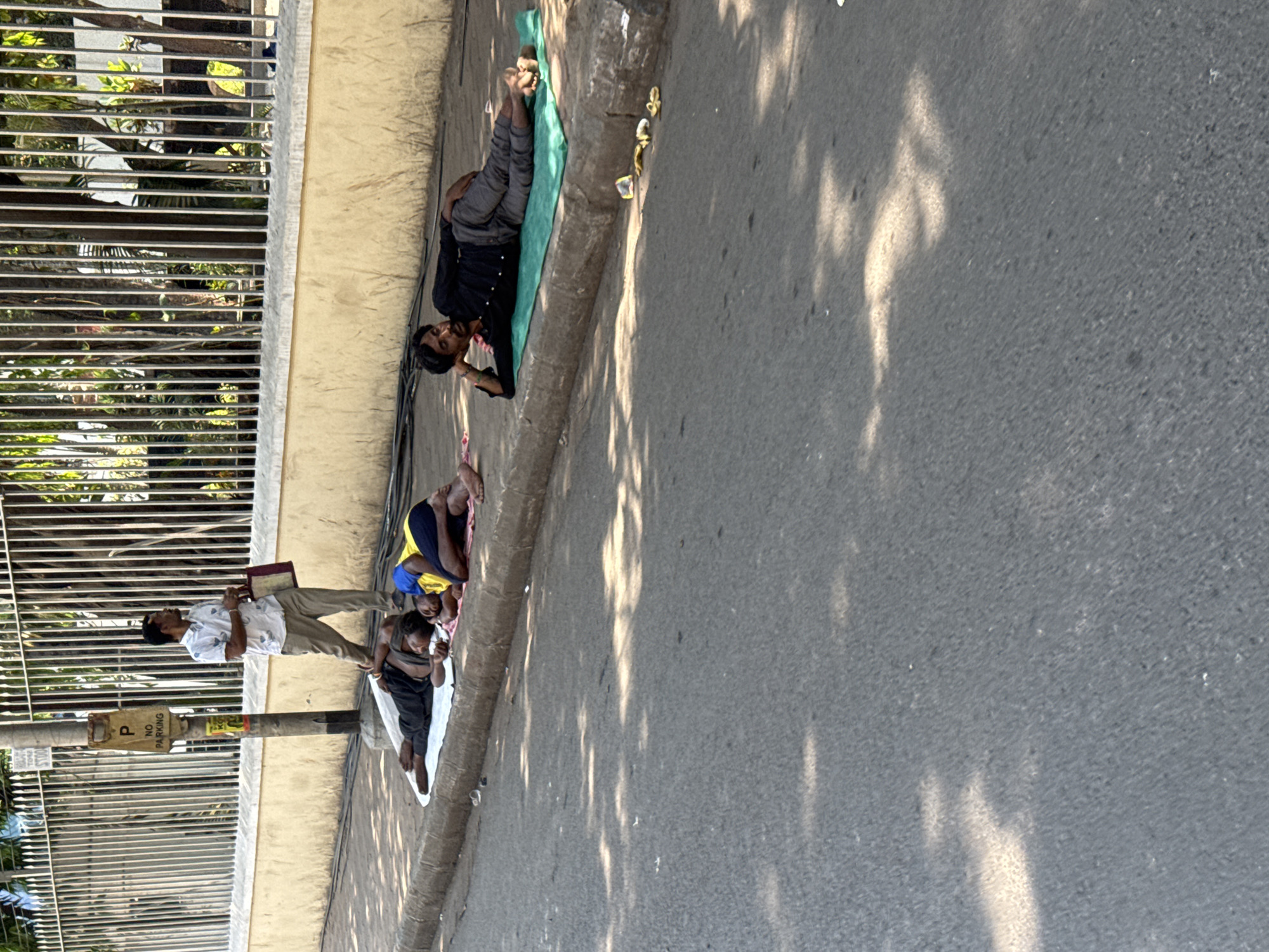 A street scene in Kolkata, India, shows several people lying on the ground next to a low wall bordering a street. One man lies on a green mat, wearing dark clothing. Two other people lie on a white sheet; one wears a blue and yellow shirt and the other a dark shirt. A man wearing a white patterned shirt and khaki pants stands holding a book or tablet near the group on the white sheet. A metal fence runs along the opposite side of the wall. A "No Parking" sign is visible on a pole next to the fence. Shadows from trees are cast across the street. Banana peels lie near the man on the green mat.