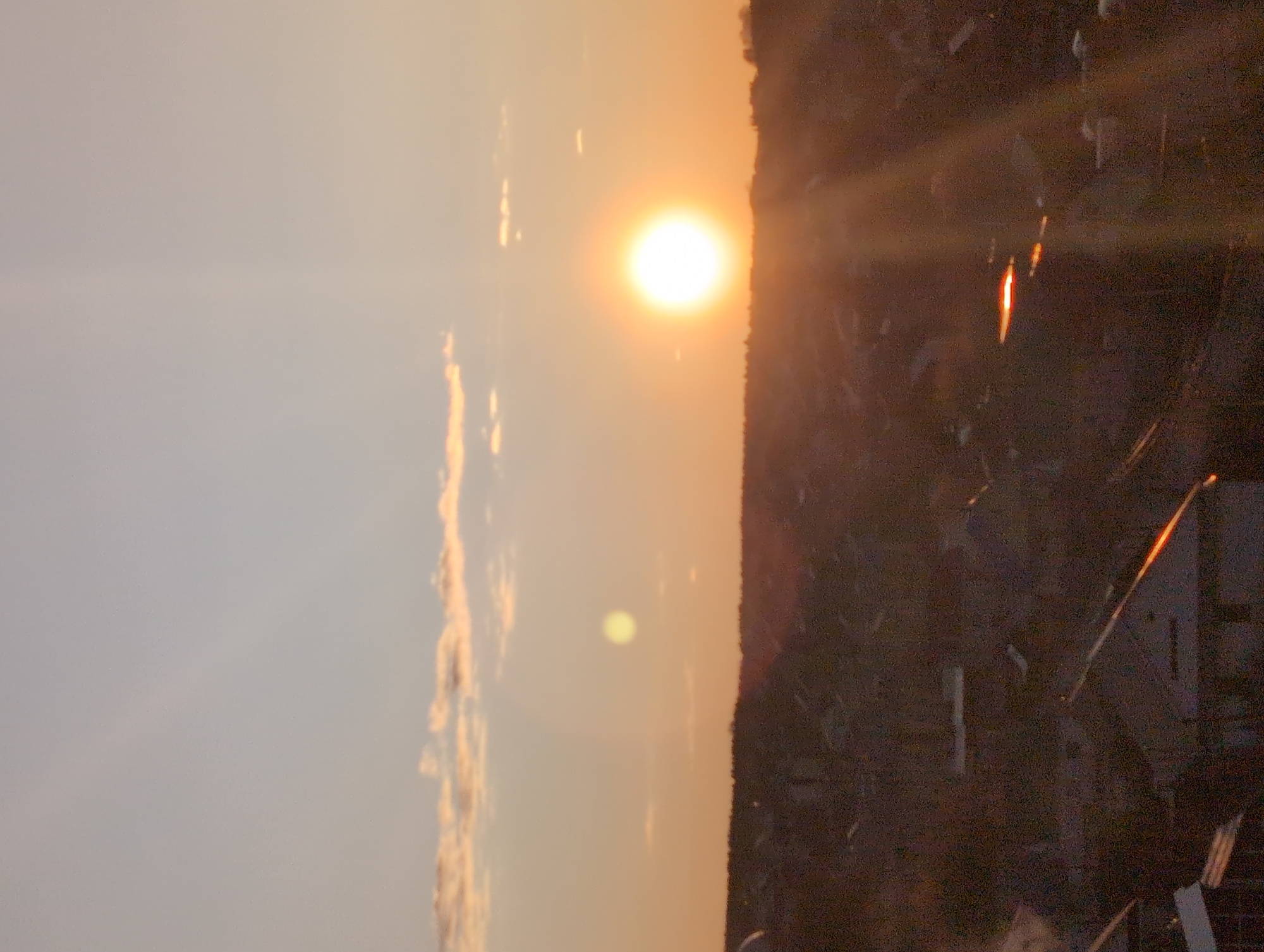 The image depicts a sunset over what appears to be a city. On the right side of the image, dark shapes suggest buildings or structures, potentially within a forested area. The sun is a bright, circular form near the top center of the frame, with rays of light extending outwards. The sky transitions from a pale blue to a warm orange near the horizon. Some clouds are faintly visible on the left side. Due to the angle, focus, and lack of identifiable landmarks, it's impossible to confirm the city is Izhevsk.