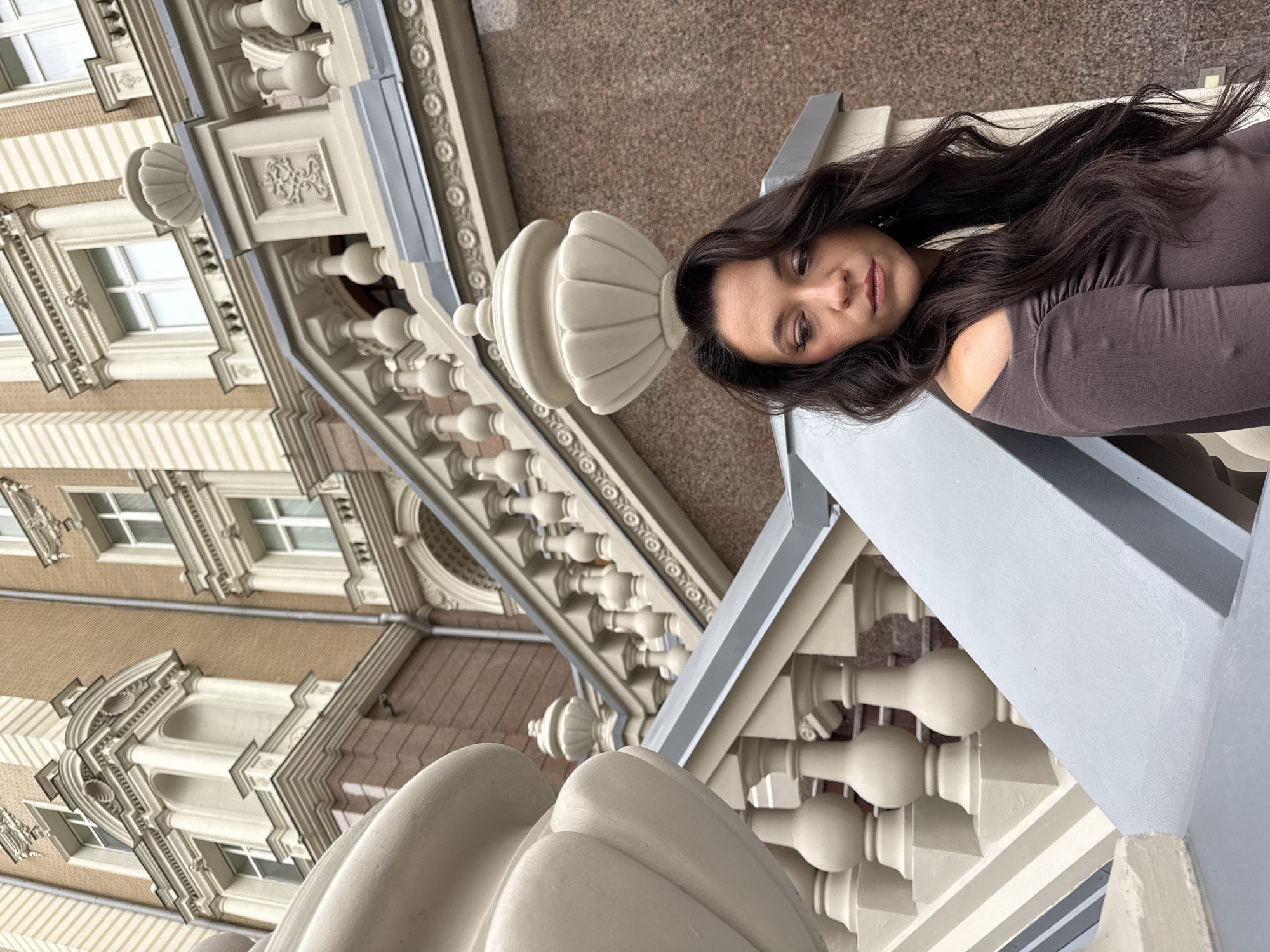 A woman with long, dark, wavy hair is standing in front of a building with ornate architecture. She is wearing a dark, off-the-shoulder top. The building features classical elements, including columns, balustrades, and decorative carvings. A grand staircase with a balustrade is prominent in the foreground and extends upwards. The woman is looking down and to her right, with her body angled away from the camera. The environment appears to be an outdoor urban setting, possibly in Kazan, Russia, given the architectural style. The lighting suggests an overcast day.