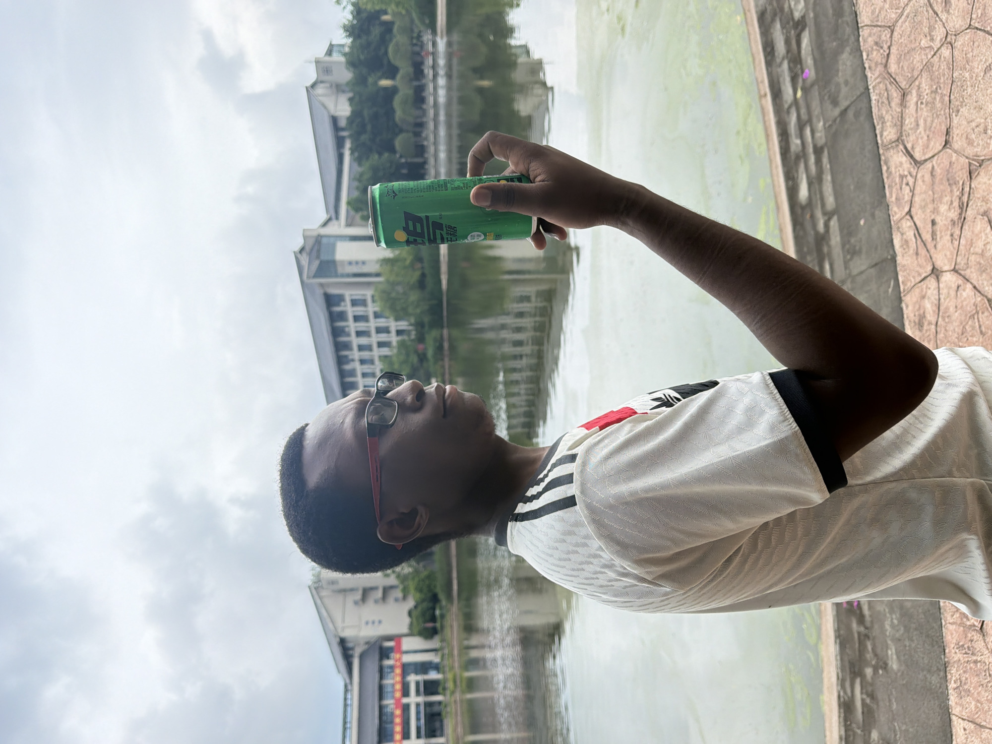 A medium shot captures a Black man in profile, wearing glasses and a white and red athletic jersey, holding a green can of what appears to be Sprite. He stands on a paved area with a textured stone pattern, overlooking a calm body of water. In the background, several modern buildings with tiered roofs and large windows are visible, set against a cloudy sky. The location is identified as Jiangjin, China. The man is holding the can up, possibly for inspection or display. The water reflects the buildings and sky. The overall scene suggests an outdoor setting, possibly a park or campus area, on an overcast day.