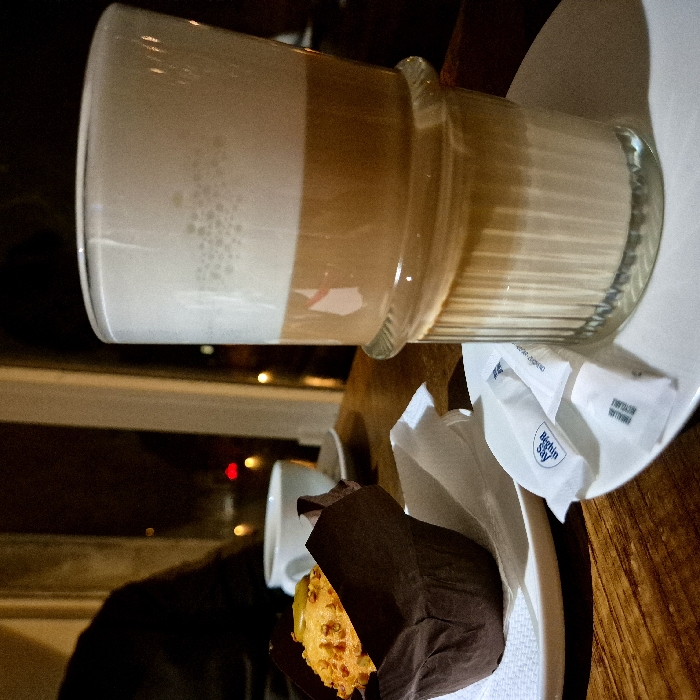 A tall glass of coffee with a thick layer of foamed milk rests on a white plate. Beside it, a muffin encased in dark brown paper sits on the same plate. Two sugar packets, one bearing the "Beghin Say" brand, are positioned near the coffee glass. The scene is set on a wooden table, reflecting ambient light from a nearby window. A white ceramic cup is visible in the background.