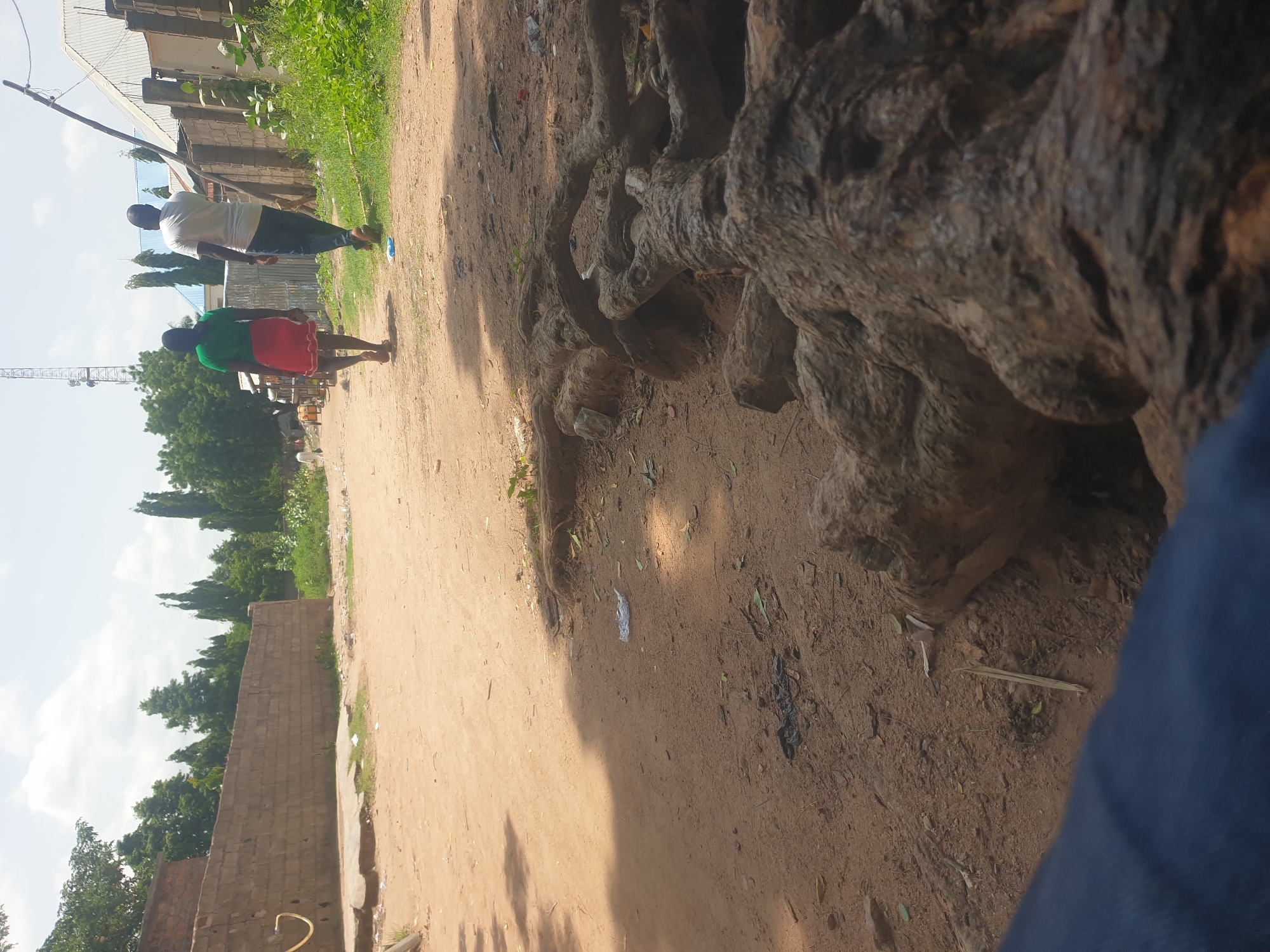 The image depicts an outdoor scene with two people walking on a dirt path. The person in front is wearing a green shirt and a red skirt, while the person behind them is wearing a white t-shirt and dark pants. They are walking away from the camera. The path is lined with trees and a brick wall on the left side. In the background, there are buildings and a construction crane. The scene appears to be set during the day, with sunlight casting shadows on the ground. The weather seems to be clear. The ground is dusty and there are some scattered pieces of debris. The overall atmosphere is casual and everyday. There is no visible text in the image.