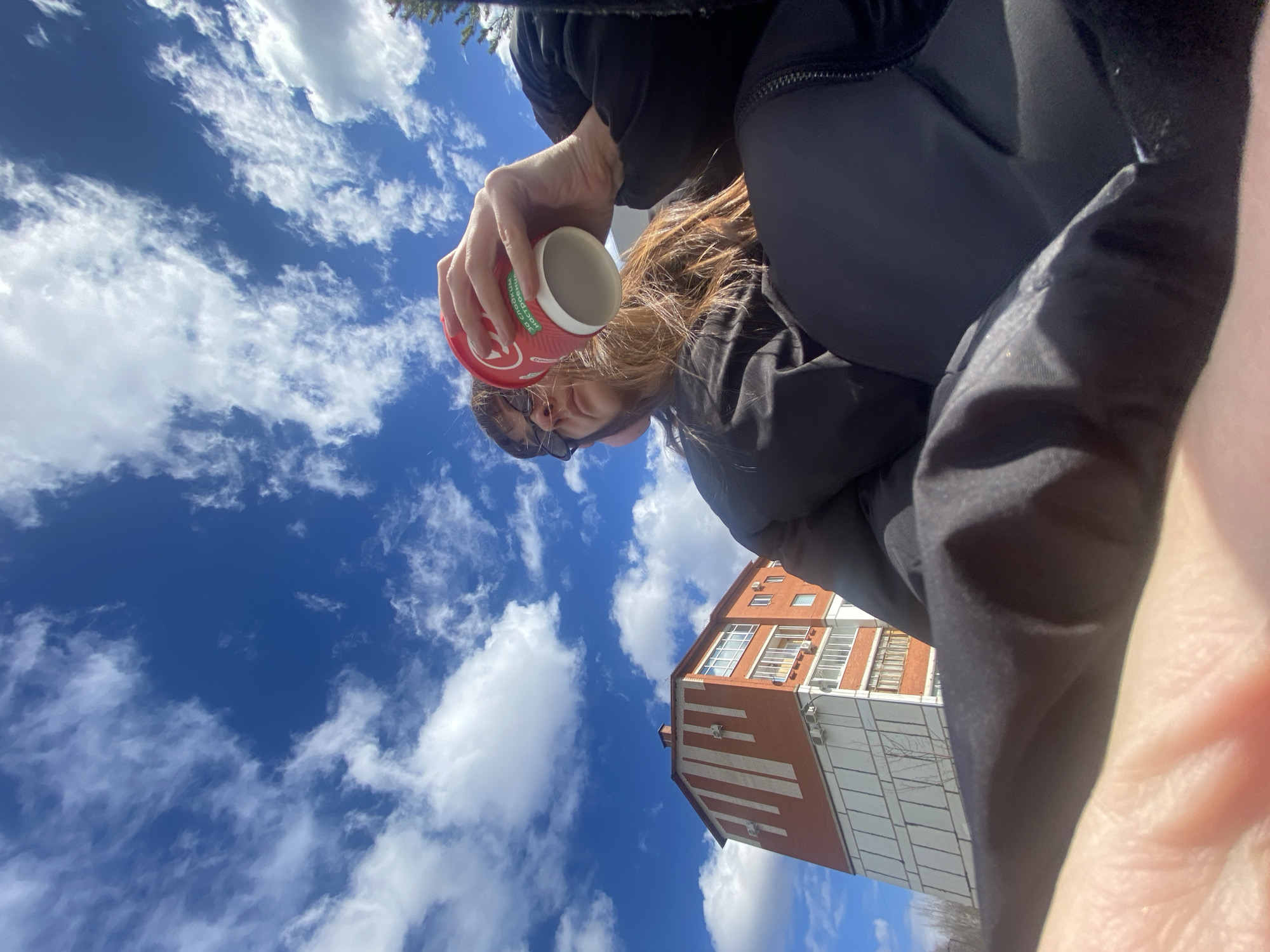 A low-angle photo shows a person holding a red and white coffee cup with the logo facing downward. The person wears sunglasses and a dark jacket. The background features a blue sky with scattered white clouds. A building with red brick and white accents is visible in the lower-right of the frame. The cup reads 'DONDO' on the side. Based on the architectural style and the 'DONDO' coffee cup logo, this scene is located in the city of Votkinsk, Russia.