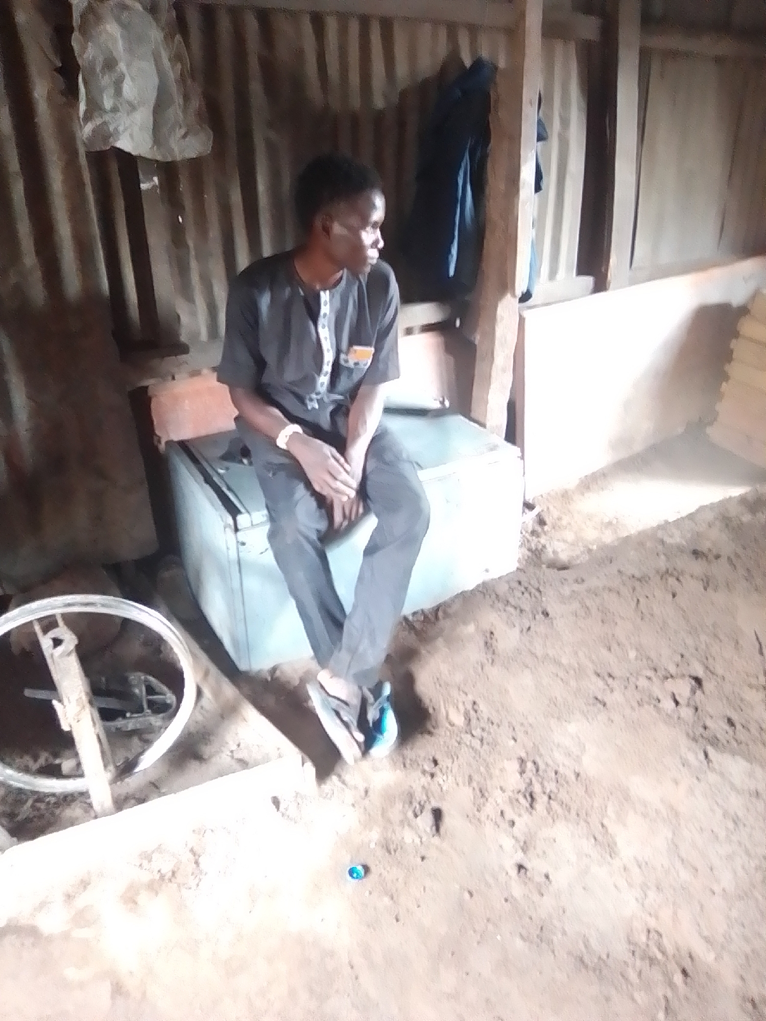 In Arawa, Nigeria, a young man wearing a dark, short-sleeved traditional outfit with white trim and a yellow badge sits on top of a light blue, rectangular metallic object. The object appears to be a refrigerator or a similar appliance, positioned on a dirt floor. His legs are crossed, and his hands rest on his lap. He is looking to his right, away from the camera. The background consists of a corrugated metal wall with wooden support beams. To the left of the man, a bicycle wheel rim is visible, resting on a wooden beam. The lighting suggests an indoor environment, possibly a workshop or a storage area, with the floor being uneven and dusty. A white plastic bag hangs from the ceiling near the corrugated wall.
