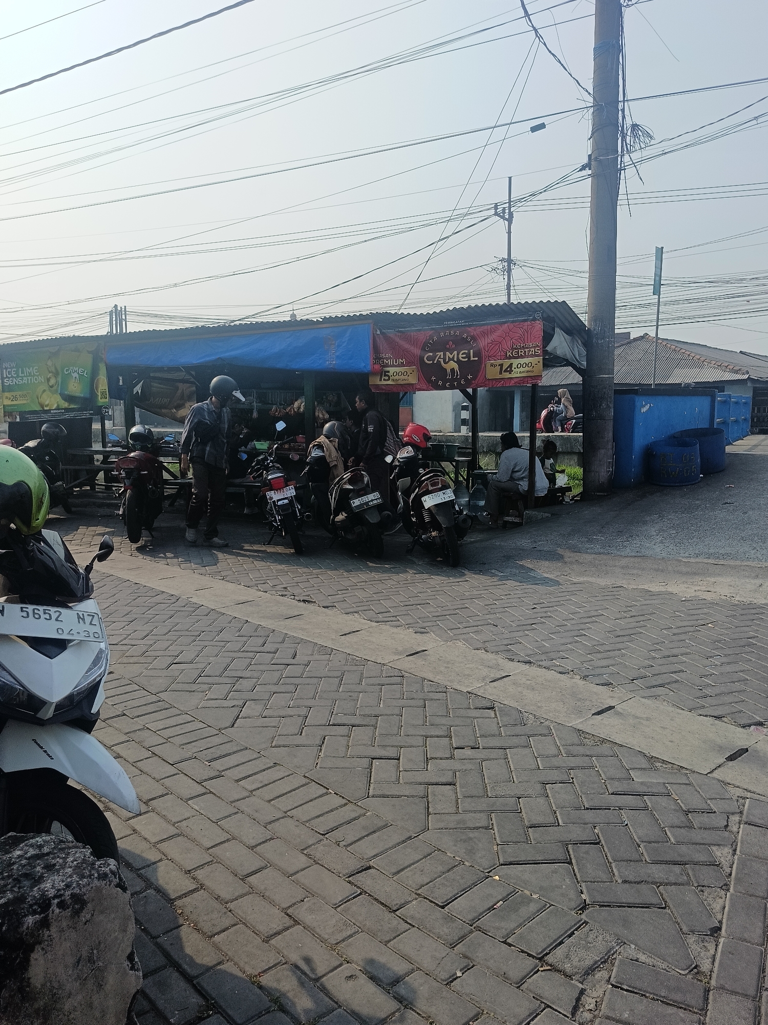 The image depicts an outdoor scene in Wedi, Indonesia, likely a street-side area with a small food or drink stall. In the foreground, a white scooter with license plate N 5652 NZ is parked on a brick-paved surface. Behind the scooter, a group of people are gathered around a covered stall. Several motorcycles are parked near the stall, some with riders wearing helmets. The stall itself has a blue tarp covering the top and is adorned with advertisements for "Camel" cigarettes, indicating prices for different packs. People are seated at tables or standing, engaged in conversation or purchasing items. In the background, blue structures, possibly more stalls or storage units, are visible, along with utility poles and overhead wires. The overall atmosphere suggests a casual gathering spot in a busy urban environment.