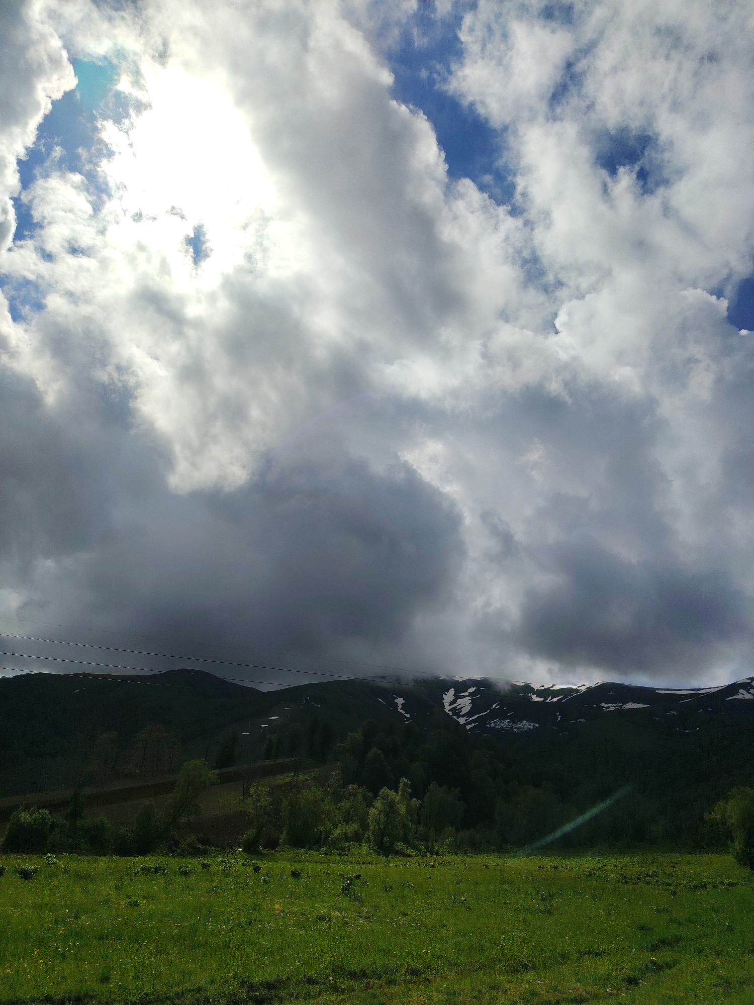 The image depicts a landscape scene with a dominant sky filled with dramatic clouds, some of which are backlit by the sun, creating bright areas. Below the sky is a dark, silhouetted mountain range. The upper portions of the mountains show patches of snow. The middle ground features a dense forest and a field that appears to be tilled or cultivated. The foreground is a vibrant green meadow with scattered yellow wildflowers. Thin, dark lines, possibly power lines or thin trees, are visible against the sky above the mountains. The overall impression is one of a natural, possibly mountainous, environment under a dynamic sky. There are no people or animals visible in the frame.