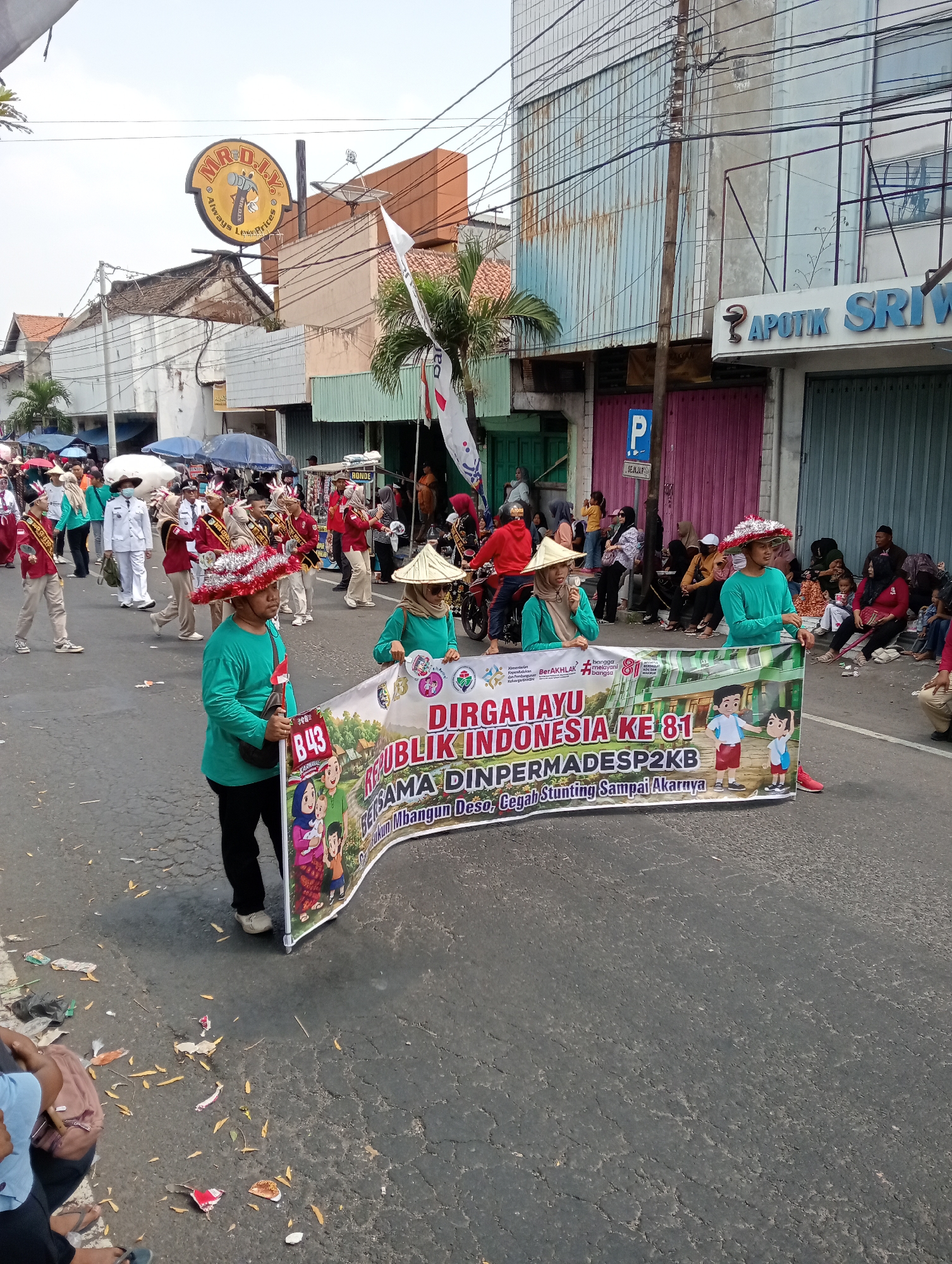 In the city of Bintoro, Indonesia, a parade is underway along a paved street lined with shops and buildings. On the right side of the street, a building with a blue and white sign reading "APOTIK SRIWIJAYA" is visible. A palm tree stands in front of the building. The street is crowded with spectators sitting on the side and standing. In the foreground, two individuals are holding a large banner that reads "DIRGAHAYU REPUBLIK INDONESIA KE 81" and features illustrations of families and children. Several other parade participants are visible, some wearing traditional hats and colorful outfits, while others are in uniform. The parade is taking place during the daytime, with overhead wires and streetlights visible. Scattered debris, including leaves and paper, litters the street.