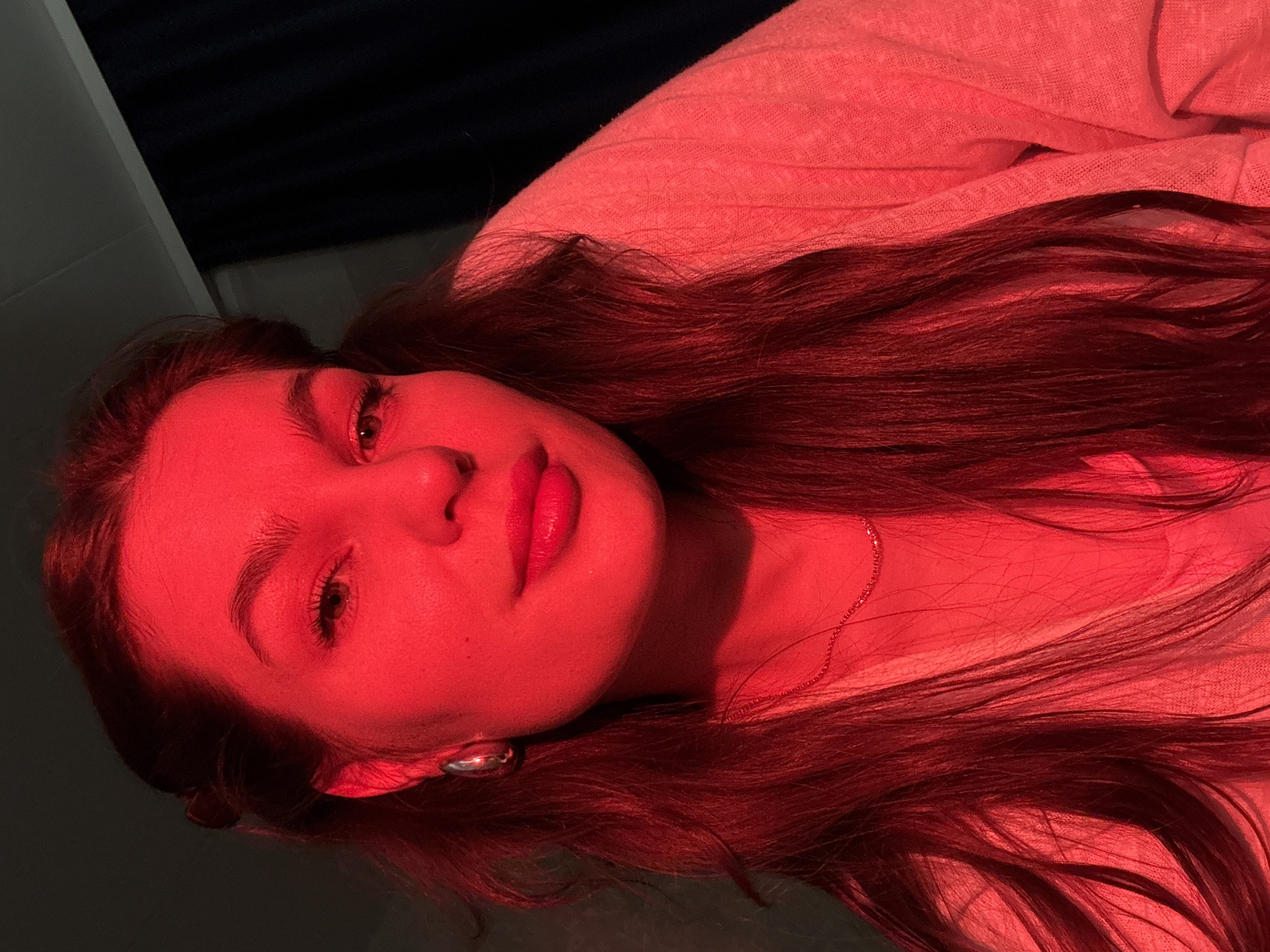 A young woman with long, dark reddish-brown hair is photographed in a room illuminated by a strong red light. She is lying down, facing the camera, with her head tilted to the side. She has fair skin, dark eyebrows, and her lips appear to be painted red. A silver earring is visible in her left ear. She is wearing a light-colored top. A thin necklace is around her neck. The background is a combination of light gray and dark, almost black, areas. The specific location within the city of Chaikovsky, Russia, cannot be determined from the given image.