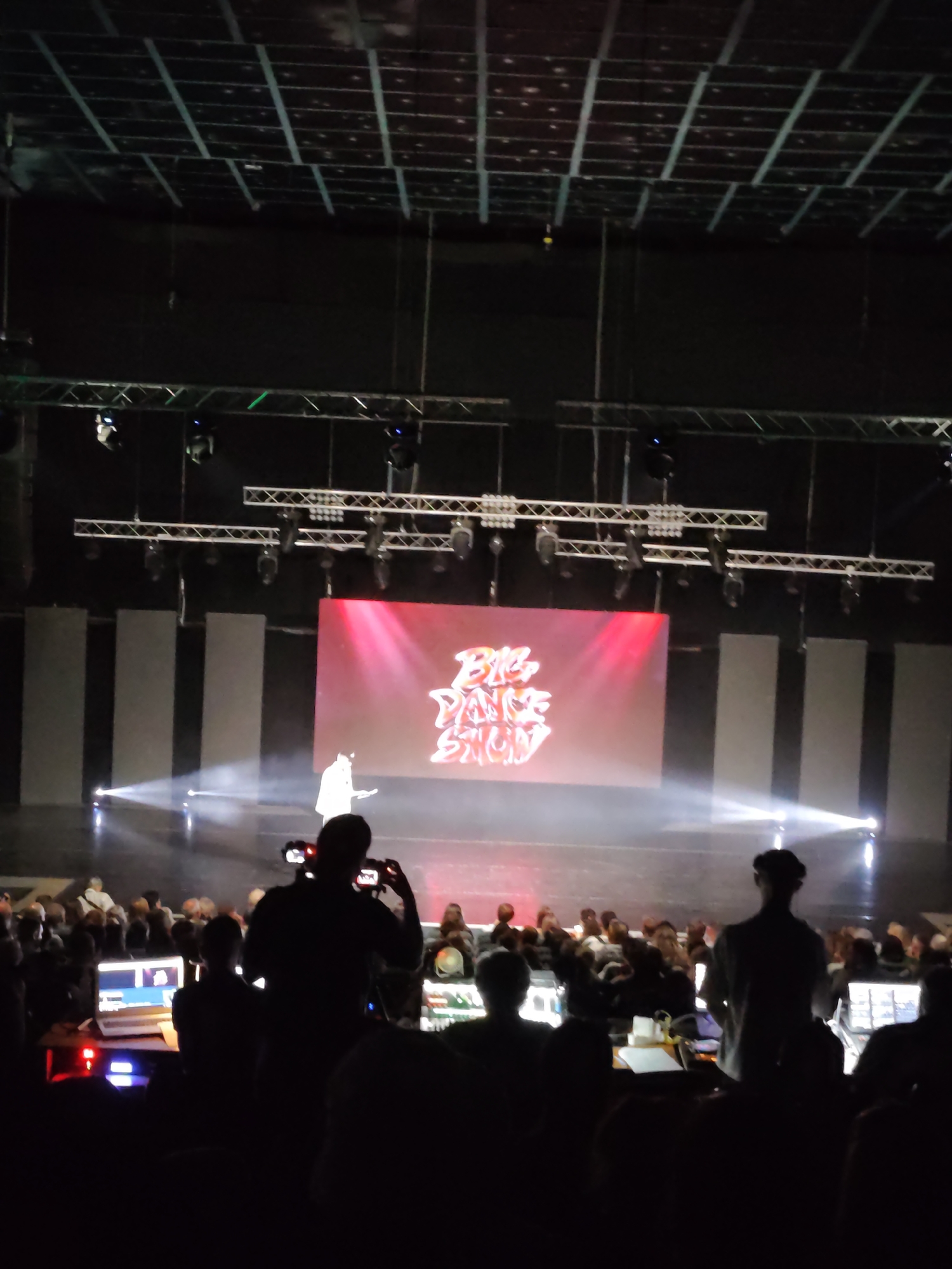 The image depicts a large indoor venue, possibly a theater or concert hall, with a stage set up for a performance. A large screen is prominently displayed on the stage, showing the words "BIG DANCE SHOW" in a stylized font, illuminated with red and pink light. A single figure, likely a performer or presenter, stands on the stage in front of the screen, bathed in white light. The audience is seated in the foreground, mostly obscured in shadow, with some individuals visible as silhouettes. In the lower part of the image, several people are seen operating technical equipment, including laptops and audio consoles, suggesting they are part of the production crew. The overall lighting is dim, with spotlights focused on the stage. The location is not specified in the image.