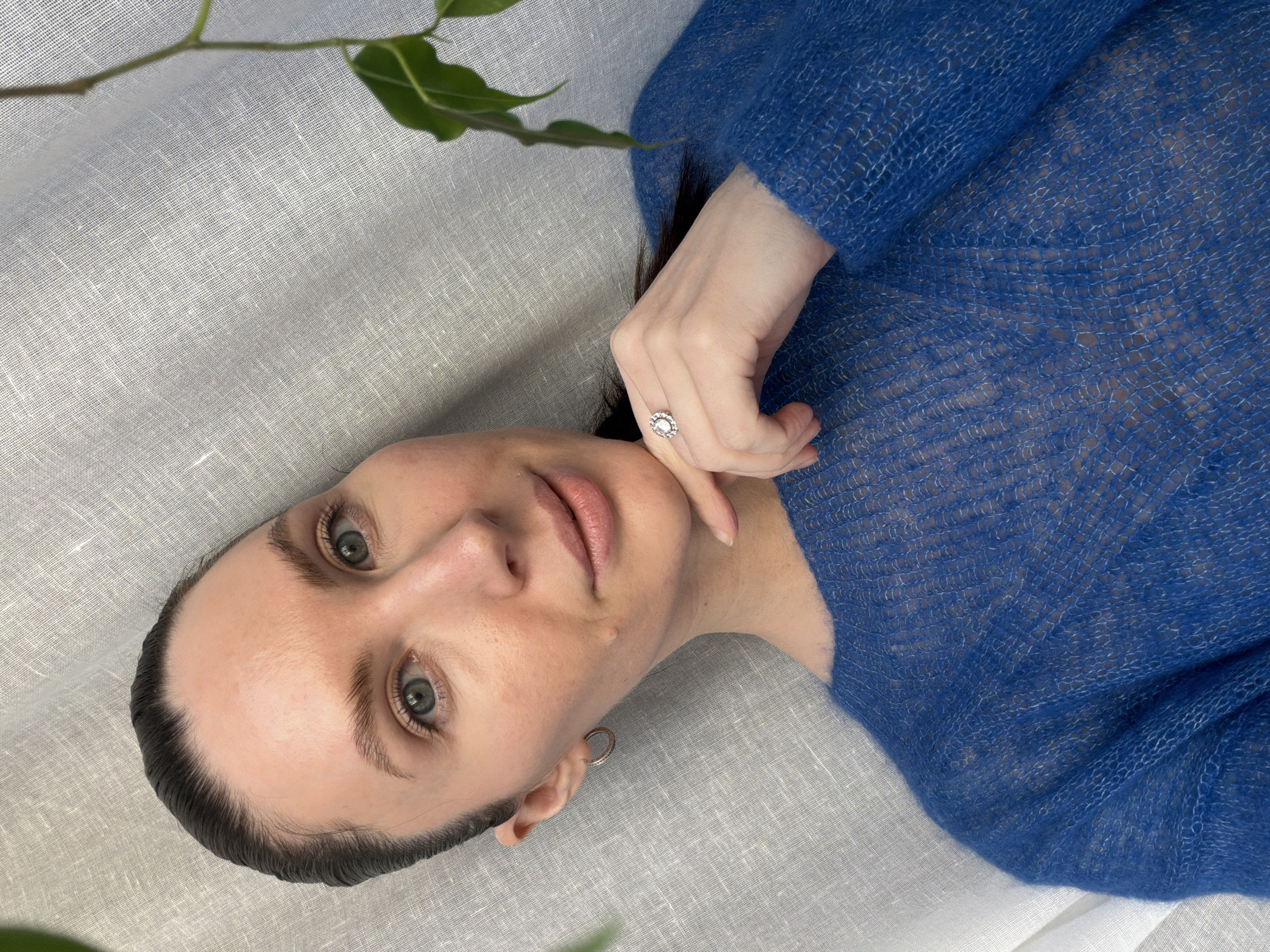 A woman lies on a textured white fabric, wearing a sheer blue sweater. Her hand, adorned with a ring, rests near her face. A leafy branch enters the frame at the top left. She wears a silver hoop earring. The photograph may have been taken in Perm, Russia, though there is no direct evidence in the image to support this.