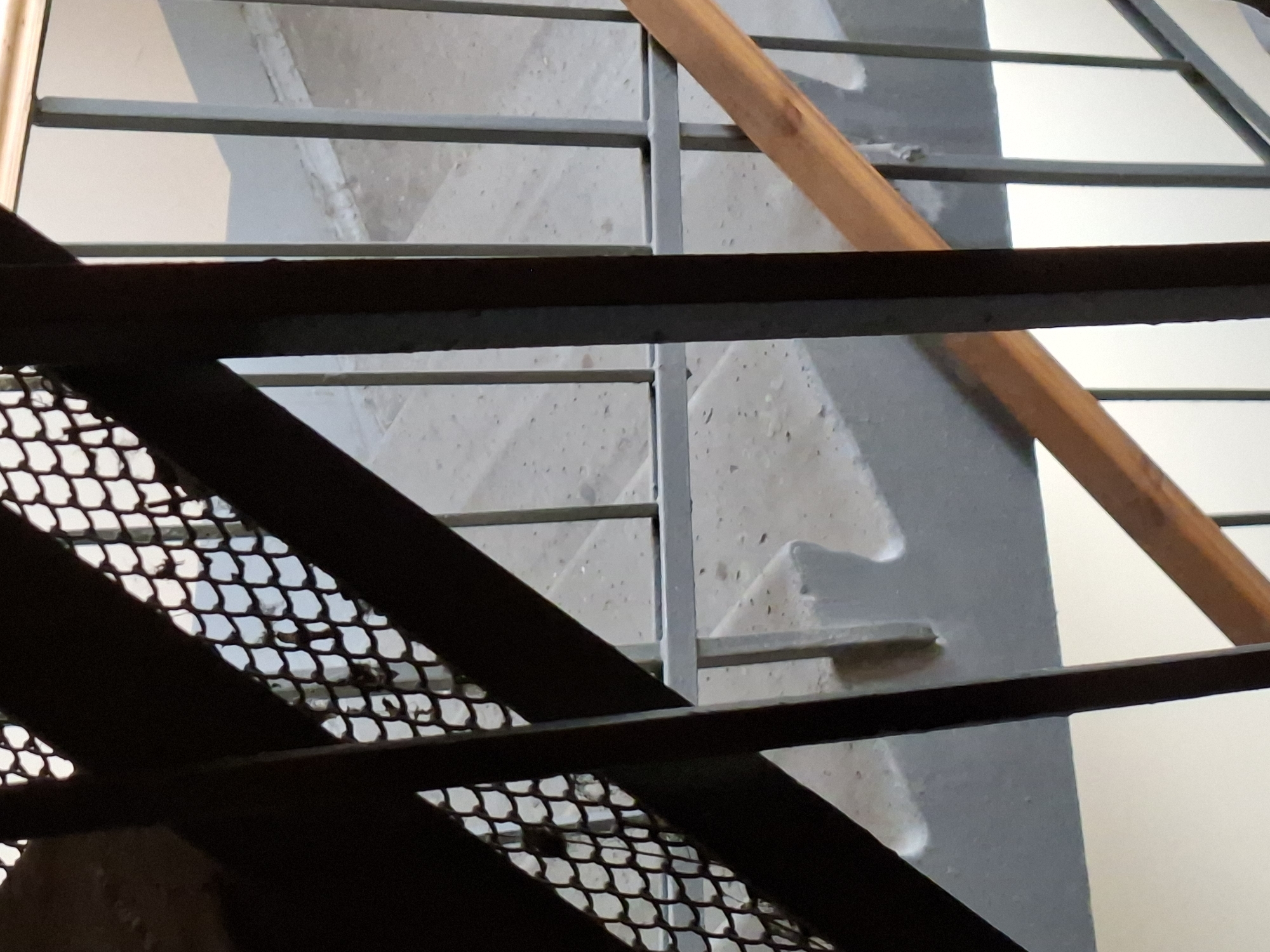 This media file is a close-up, abstract photograph of a section of an outdoor staircase, likely in Kazan, Russia. The primary subjects are the architectural elements of the staircase itself: metal railings, metal mesh grating, and concrete steps. A wooden beam runs diagonally through the upper portion of the frame, adding a contrasting texture and color.
The image captures a static scene, with no people or overt activity. The focus is on the interplay of light and shadow, the geometric lines of the structure, and the rough texture of the concrete. The time of day and weather are not explicitly discernible, though the lighting suggests it could be daytime, with light casting shadows on the concrete.
Notable details include the fine mesh of the grating, which allows a glimpse of the background, and the smooth, angled surface of the concrete steps. The metal components appear to be made of dark, possibly rusted metal, contrasting with lighter grey or white painted surfaces. The wooden beam has a natural, grainy texture and a warm, reddish-brown hue.
The overall impression is one of urban infrastructure, possibly an industrial setting or a functional outdoor space. The image does not contain any visible text. The abstract nature of the composition, with its cropped view and focus on form and texture, invites the viewer to appreciate the aesthetic qualities of everyday structures.
