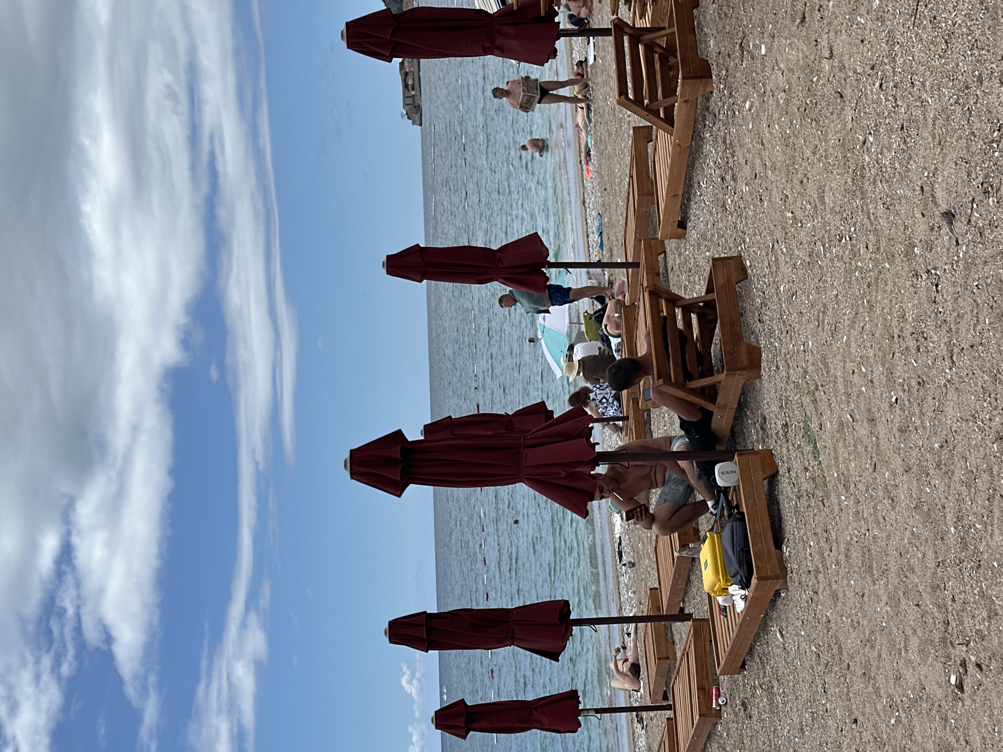 This image captures a vibrant beach scene in Budva, Montenegro, on a clear, sunny day. The perspective is from the sandy shore, looking out towards the sea and sky, though the image is rotated 90 degrees clockwise.
In the foreground, several dark red, folded beach umbrellas are arranged in a row, casting light shadows on the sand. Beside them are wooden beach loungers, some occupied by people relaxing. One individual in the mid-ground, seated on a lounger, is holding a phone and has a cigarette in their mouth. Nearby, a yellow bag and a black bag rest on or near a lounger, along with a white portable speaker or device clearly labeled "SOUTH".
Further towards the water, several people are visible. One person stands in the shallow water, engrossed in reading a newspaper. Another person is partially submerged in the sea. In the background, the calm, blue-green sea stretches out, dotted with what appear to be small buoys or markers. The horizon line is visible, beyond which a hazy landmass or distant coastline can be faintly seen.
The sky is a bright, clear blue with streaks of wispy white clouds, indicating fair weather. The overall atmosphere suggests a leisurely summer day at the beach, with people enjoying the sun and water.