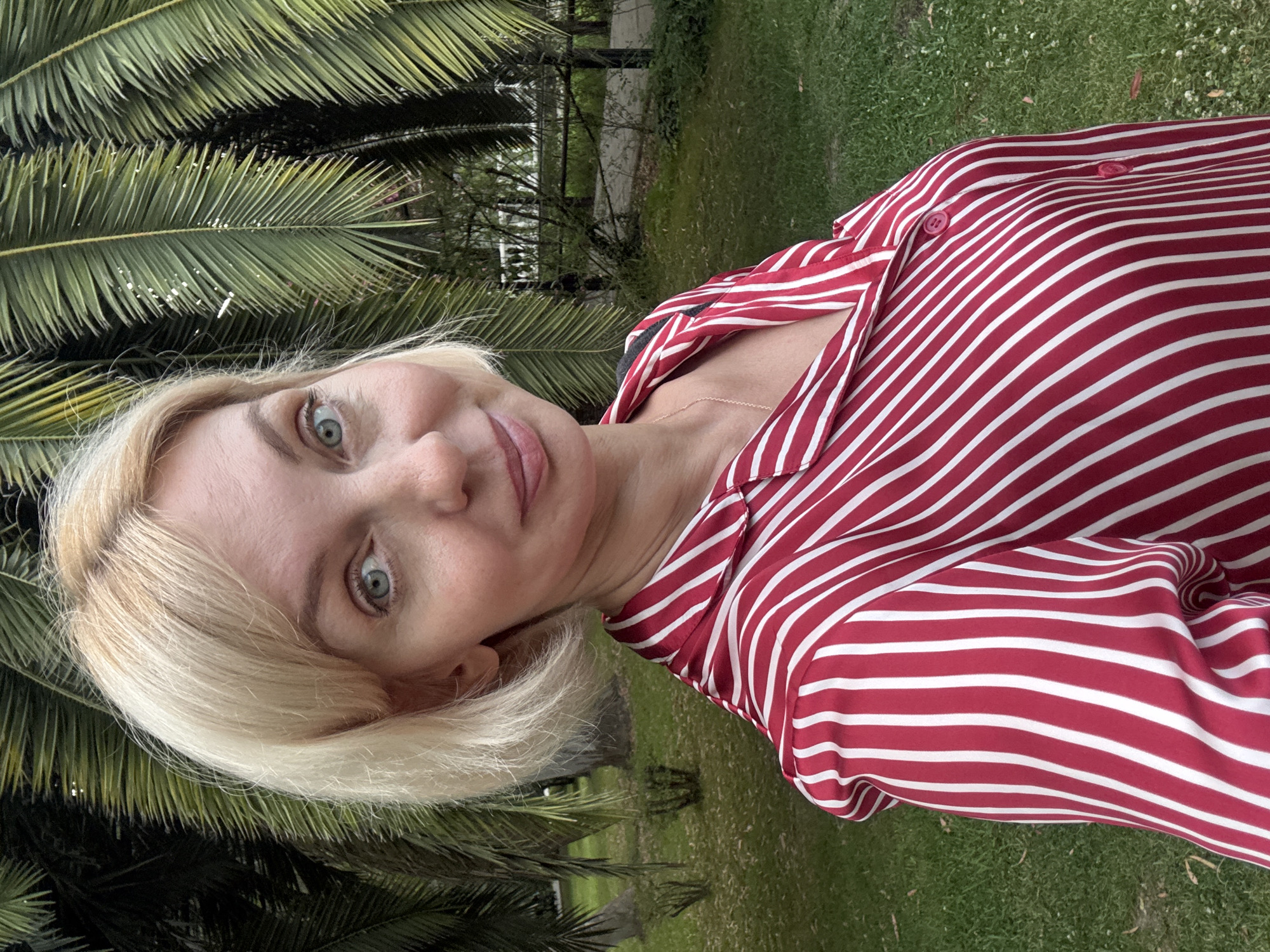 The media file shows a close-up, sideways selfie of a woman with blonde hair and blue eyes. She is wearing a red and white striped shirt. Her head is tilted, and she is looking towards the camera with a slight smile. Behind her, palm fronds are visible, suggesting a tropical or subtropical setting. The background also includes green grass and some blurred foliage, possibly indicating an outdoor location like a park or garden. The lighting suggests it is daytime, with no specific indication of time or weather conditions. The image appears to be taken in Sochi, Russia, a location known for its subtropical climate and palm trees. There is no visible text in the image.