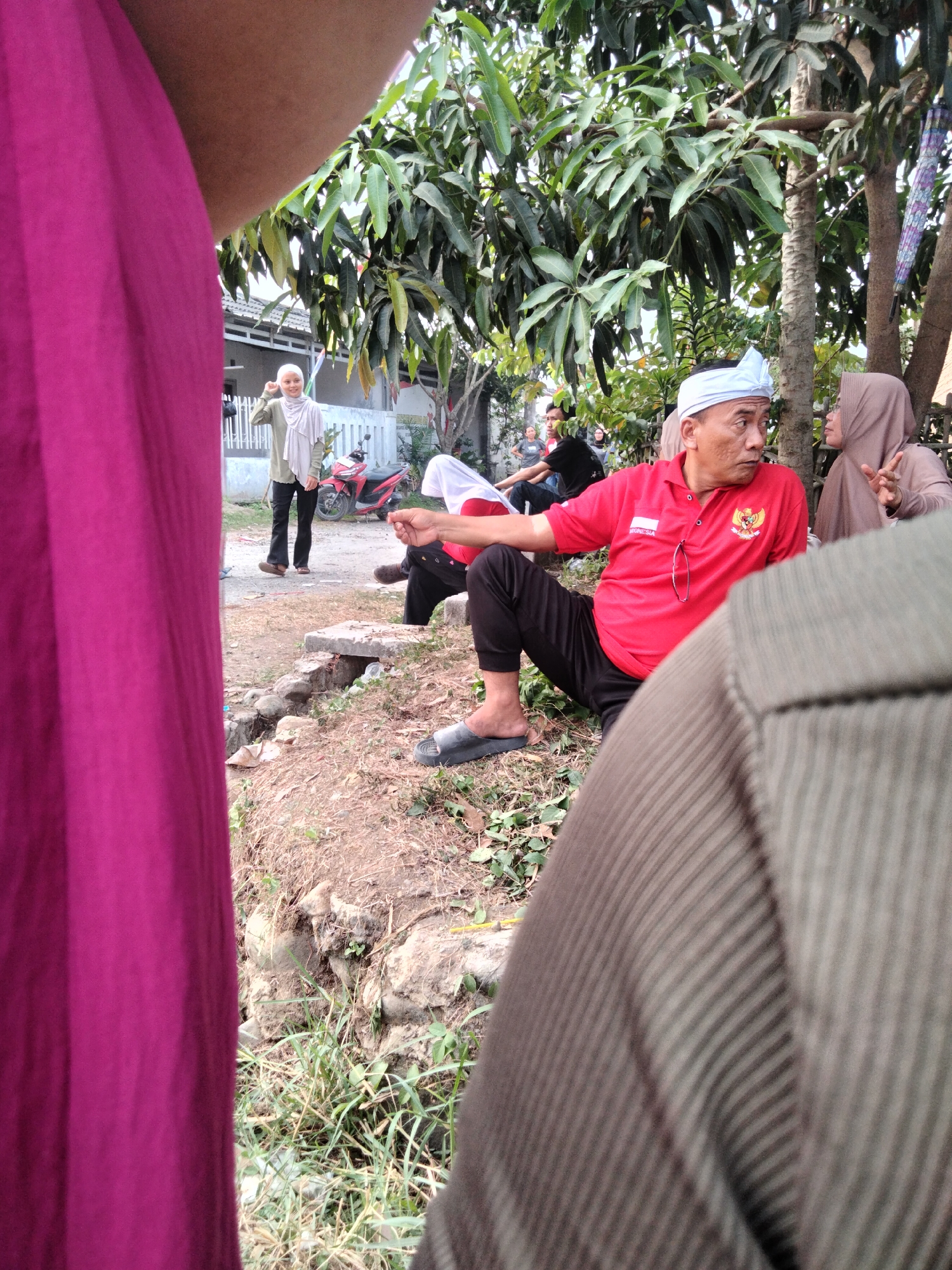 In the city of Cariu, Indonesia, a group of people are gathered outdoors. In the foreground, a person wearing a magenta garment is partially visible. To the right, another individual is covered by a textured brown fabric. In the middle ground, a man in a red polo shirt and black pants, wearing a white head covering, is seated and looking to his right. He has glasses hanging from his shirt. To his right, a woman in a beige hijab is also seated and gesturing with her hands, appearing to be in conversation with the man. Behind them, other individuals can be seen in the background, including a woman in a light green hijab standing near a red motorcycle. The environment is a dirt path with some greenery and trees overhead.