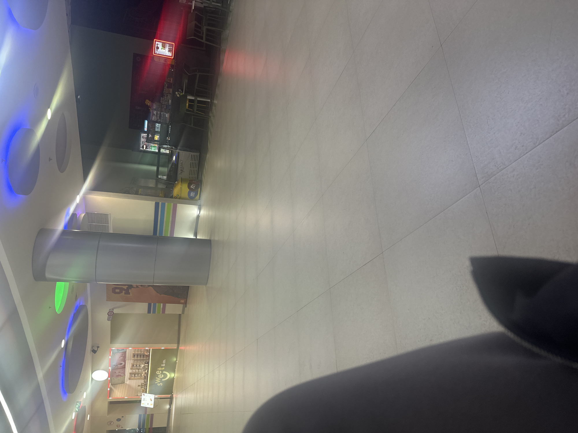 The image depicts an interior space with a polished tile floor. In the background, a food court area is visible with signs and seating. A large, cylindrical support column stands in the foreground, near a storefront. The view is slightly obstructed by what appears to be a dark piece of clothing. The location appears to be a shopping mall in Yerevan, Armenia.