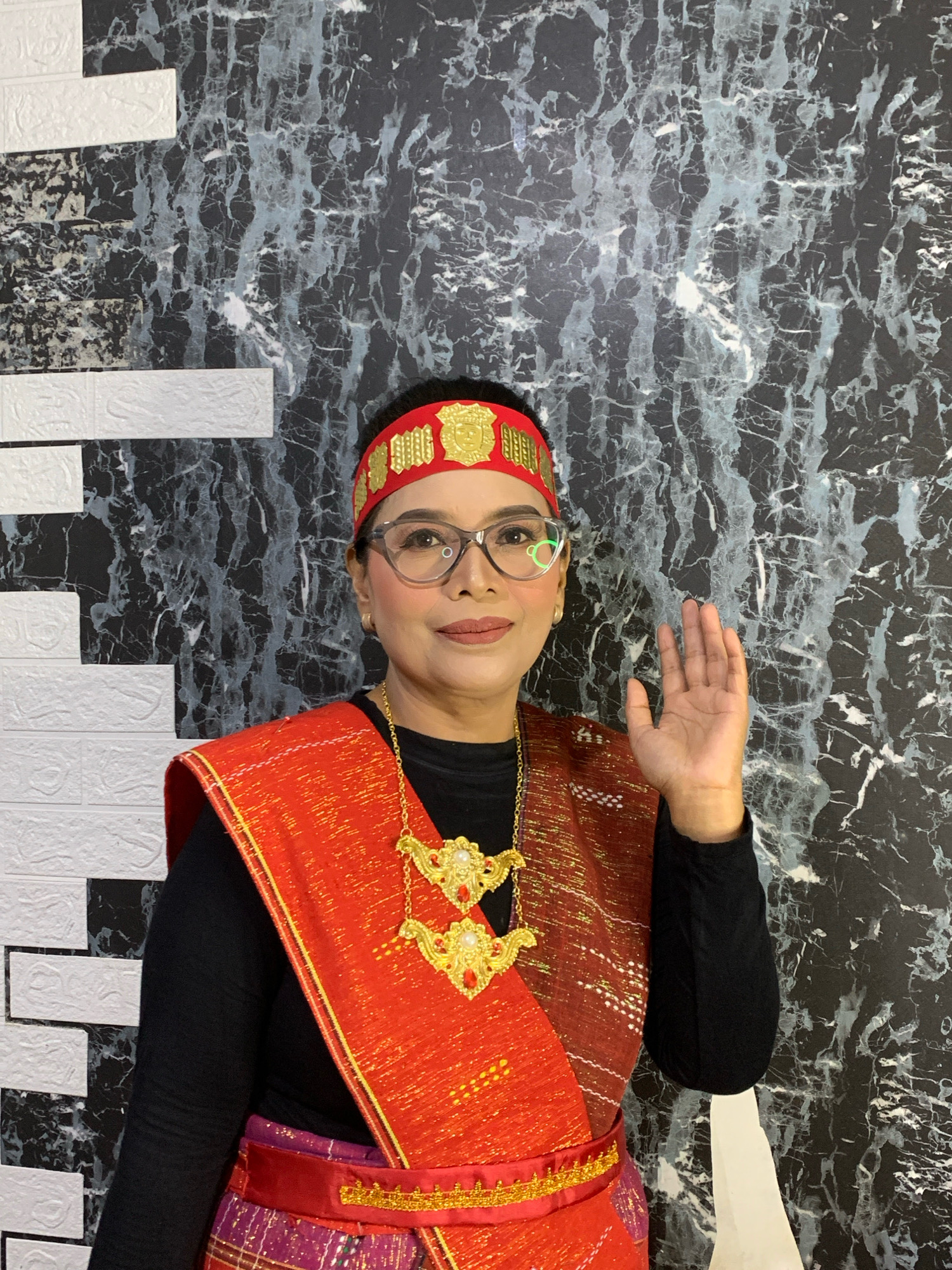 A medium shot shows a middle-aged Indonesian woman, likely in her 40s or 50s, standing and posing for a photograph in front of a dark, marble-patterned wall. She is wearing traditional clothing, featuring a bright red sash with gold accents draped over a black long-sleeved shirt, and a vibrant, patterned skirt. A red and gold headband adorns her forehead, and a striking gold necklace with red embellishments hangs around her neck. She is wearing glasses and has makeup on, with her right hand raised in a greeting gesture. The background wall has a textured, black and grey marbled pattern, with sections of white textured material resembling bricks on the left side. The lighting is bright, illuminating the woman and her attire. The overall scene appears to be a studio portrait or a staged photograph taken indoors, possibly in a residential setting or a place for cultural events in the city of Malang, Indonesia.