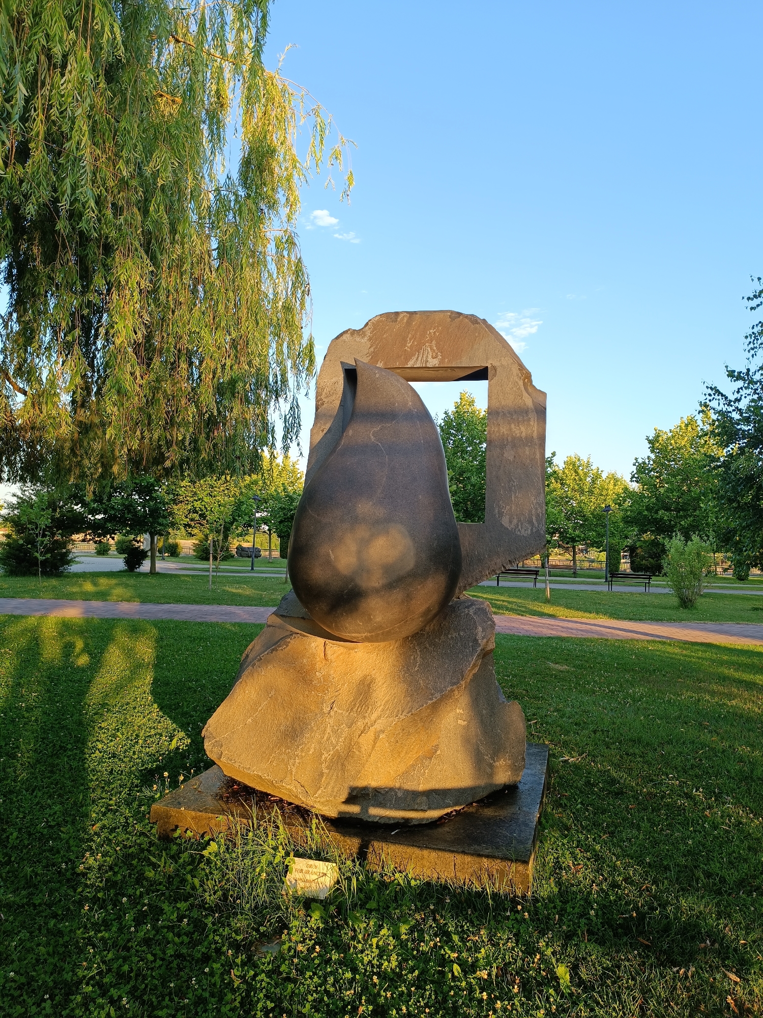 The image depicts a contemporary stone sculpture situated in a park setting. The sculpture consists of two main components: a large, dark, teardrop-shaped element resting on a rough, multi-faceted stone base, and a stylized, abstract stone archway that partially frames the teardrop. The sculpture is placed on a dark, square plinth, which is surrounded by green grass. A small, weathered sign is partially visible at the base of the sculpture, near the grass.
The park environment includes a pathway paved with red bricks winding through the grass, and several mature trees with lush green foliage. In the background, more trees line a distant area, and park benches are visible. The sky is a clear, light blue with a few wispy white clouds. The lighting suggests it is either early morning or late afternoon, with the sun casting long shadows across the lawn.
This sculpture is part of the ensemble of Brancusi's works in Târgu Jiu, Romania. Specifically, it is identified as "The Kiss" (Kissing Gate), also known as "Poarta Sarutului."