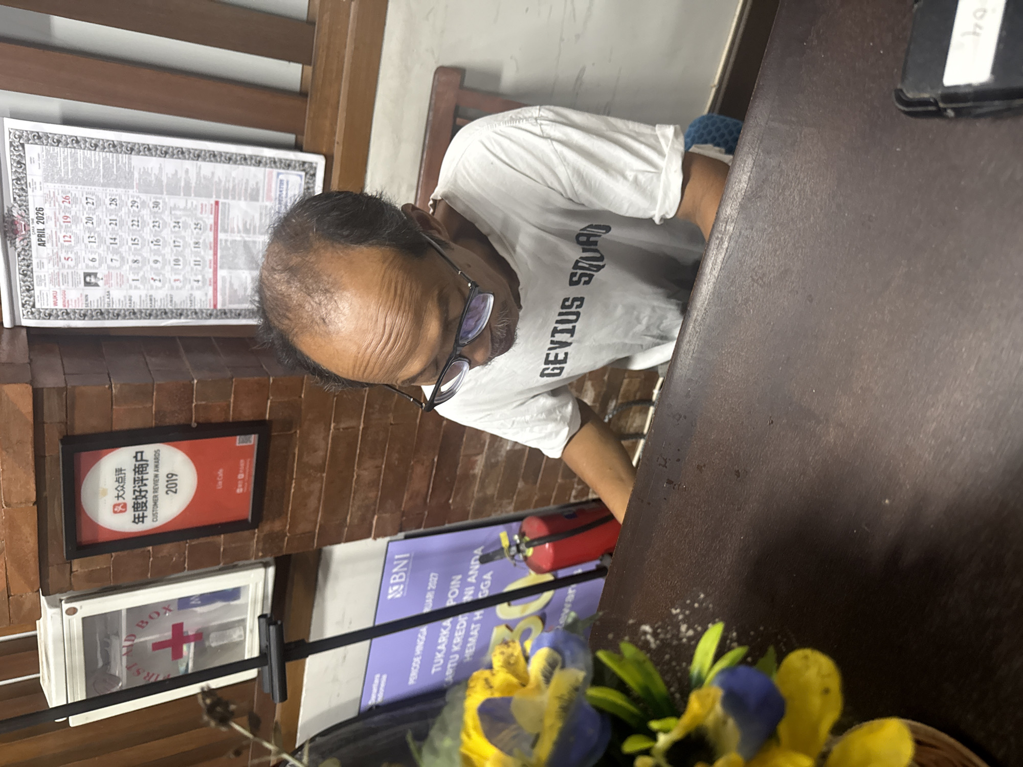 The image depicts an interior scene, likely a restaurant or cafe in Kuta Selatan, Indonesia. A man wearing glasses and a white t-shirt with the text "GEVIUS SQUAD" is seen at a table. Behind him, on the wall, is a framed calendar of April 2026, a customer review award from "大众点评" (Dàzhòng Diǎnpíng) for 2019, a first aid box, and an advertisement banner from BNI. There is also a red fire extinguisher. Yellow and blue flowers are placed in the foreground, partially obscuring the table. The table is dark brown. A dark device is on the table's corner.