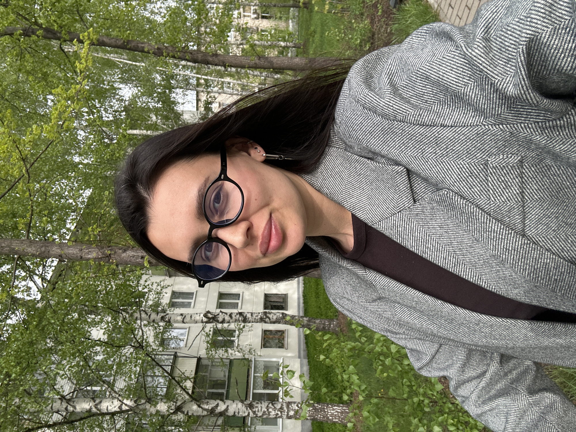 A woman with long dark hair, wearing round black glasses and a grey herringbone blazer over a dark brown shirt, is taking a selfie outdoors. Behind her is a birch tree and a multi-story apartment building with visible windows. Green trees are also present in the background and the location is likely in a park or green area near a residential building in Chaikovsky, Russia.