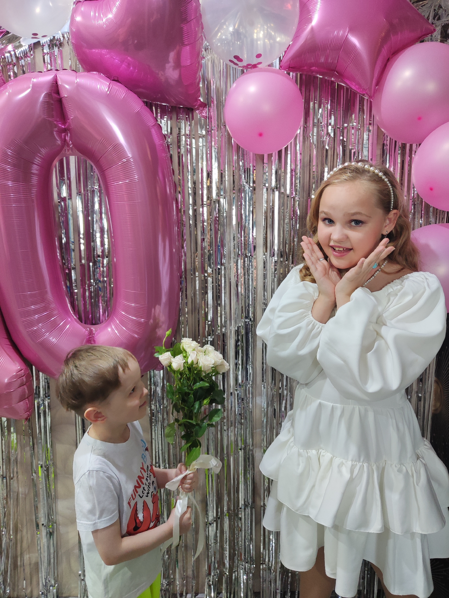 This media file captures a festive indoor scene in Chaikovsky, Russia, featuring two children at what appears to be a birthday celebration. The backdrop consists of a shimmering silver tinsel curtain, adorned with various balloons, including large pink foil number balloons. A prominent pink "0" is visible, suggesting an age like "10" or "20." Smaller pink round balloons and some white clear balloons (some with confetti) complete the cheerful decor.
In the foreground, a girl and a boy are interacting. The girl, who appears to be celebrating, stands on the right, striking a playful pose with her hands cupped around her face, smiling broadly. She has blonde hair styled with a pearl headband and is wearing a white off-the-shoulder dress with voluminous puffed sleeves and a tiered, ruffled skirt. She also wears a pearl necklace and rings.
On the left, a younger boy, with light brown hair, is presenting a bouquet of white roses, tied with a white ribbon, to the girl. He is looking up at her with a gentle smile. He wears a white short-sleeved t-shirt with a graphic design that includes what looks like a red crab and partially visible text, "FUNNY B..." and "LET'S SWIM WITH...", paired with neon green shorts.
The overall atmosphere is one of joy and celebration, marked by the bright decorations and the affectionate exchange between the children. The time of day is not explicitly indicated but appears to be well-lit indoors.