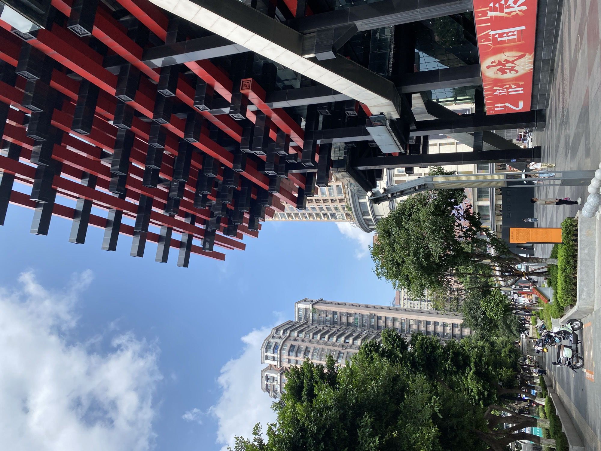 This media file shows an outdoor scene in Yuzhong District, China, likely during the daytime under a clear blue sky with some clouds. The image is captured from a low angle, looking upwards towards a modern architectural structure.
The foreground features a prominent architectural element composed of intersecting red and black beams, possibly a roof or canopy, with square light fixtures attached to the underside of the black beams. Beyond this structure, taller buildings with numerous windows rise into the sky. Some of these buildings appear to be residential apartments, characterized by their layered balconies. Lush green trees line a walkway or street in front of the buildings, adding a natural element to the urban landscape.
In the background, several people are visible, some walking along the street and others gathered near parked vehicles, which include motorcycles and electric scooters. The presence of these vehicles suggests a lively urban environment. There is a large red banner with Chinese text hanging from one of the buildings. The text appears to be in a decorative font and is the primary visible text in the image. The overall scene conveys a sense of a bustling city with a blend of modern architecture and natural elements.