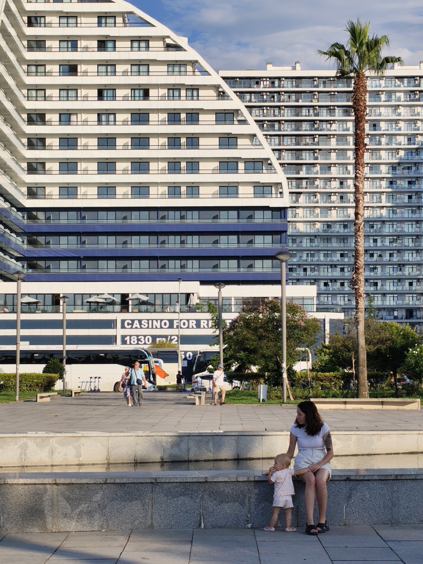 A woman and a toddler are sitting on a granite ledge in a public plaza. The woman is wearing a white t-shirt and white shorts, and the toddler is wearing a pink onesie. Behind them, a large, modern building with a distinctive geometric design, reminiscent of a stepped pyramid, is visible. This building is situated in Batumi, Georgia, and its architecture is characteristic of the city's contemporary urban development. A tall palm tree stands in front of another large building, also part of the cityscape. In the background, there are buses, people walking, and trees, suggesting a lively urban environment. The lighting indicates it is daytime, likely afternoon, with the sun casting shadows.