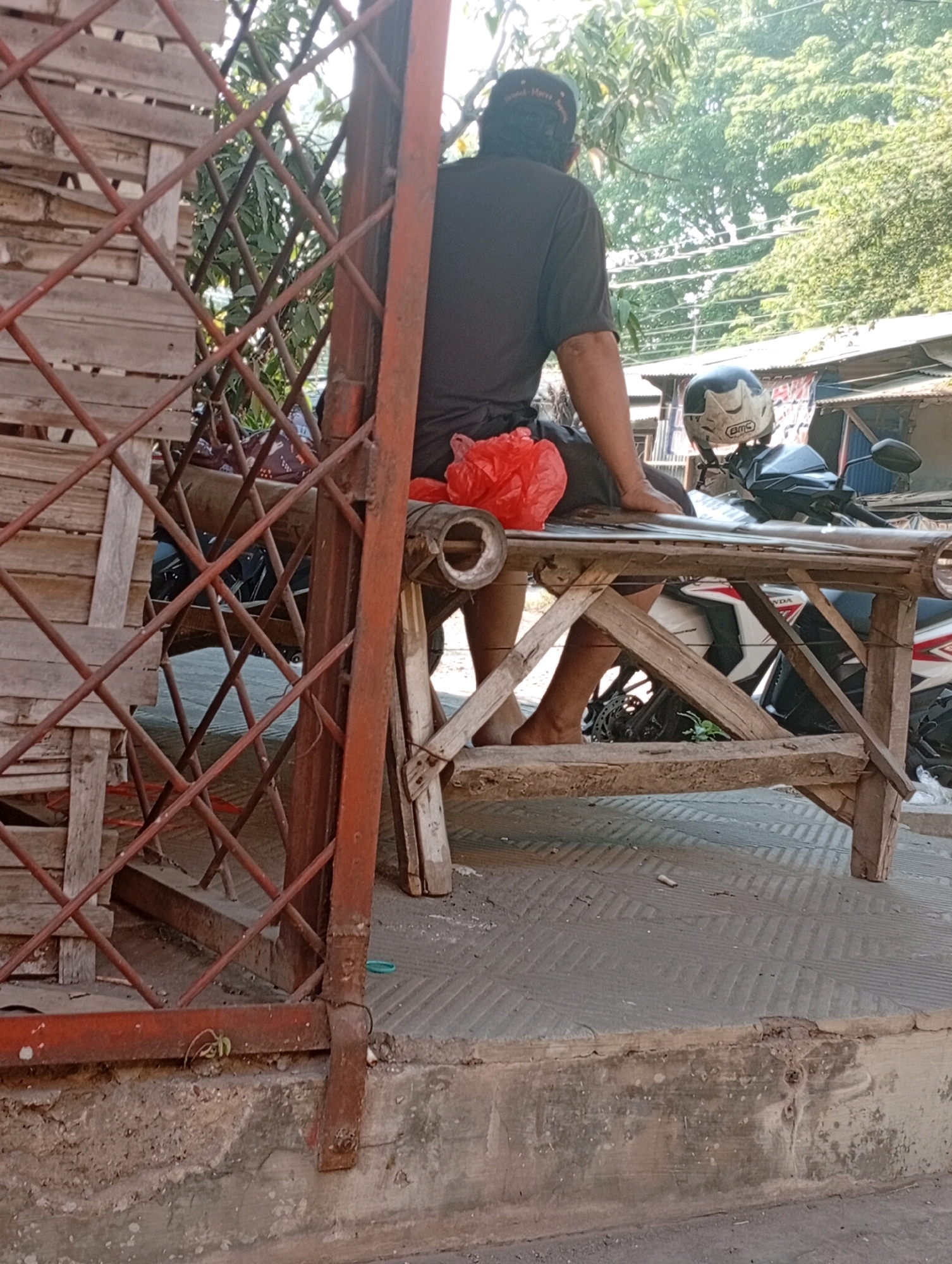 The image shows a man wearing a dark t-shirt and a dark baseball cap, seated on a wooden structure, possibly a bench or a makeshift table, outdoors. He is partially obscured by a rusty metal gate. To his left, a red plastic bag is placed on the wooden surface. In front of him, a motorcycle is parked, with a helmet resting on its front. The background reveals lush green trees and some buildings with corrugated metal roofs, suggesting an urban or semi-urban environment. The ground is paved with patterned tiles, and some debris, including a cigarette butt, is visible on it. A small plant is growing near the base of the metal gate. The lighting indicates it is daytime, likely sunny, casting shadows. The scene appears to be in Jogosari, Indonesia, as indicated by the context.
