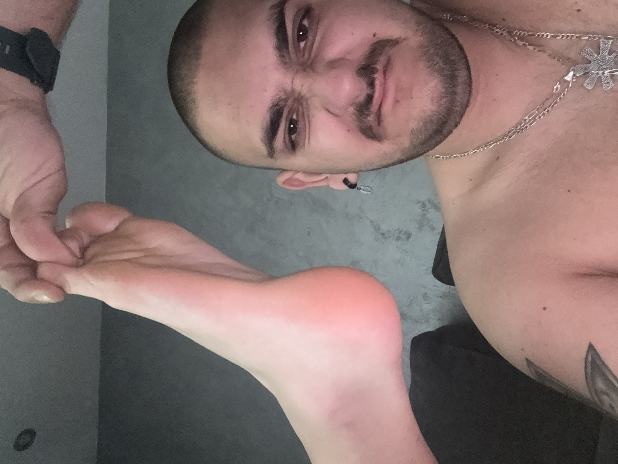 A shirtless man with a shaved head, a beard, and a silver chain is holding his foot in his hand. He is wearing a watch on his left wrist. The background is a plain gray wall. The man has a piercing in his left ear and a tattoo on his right arm. The location is not discernible from the image.