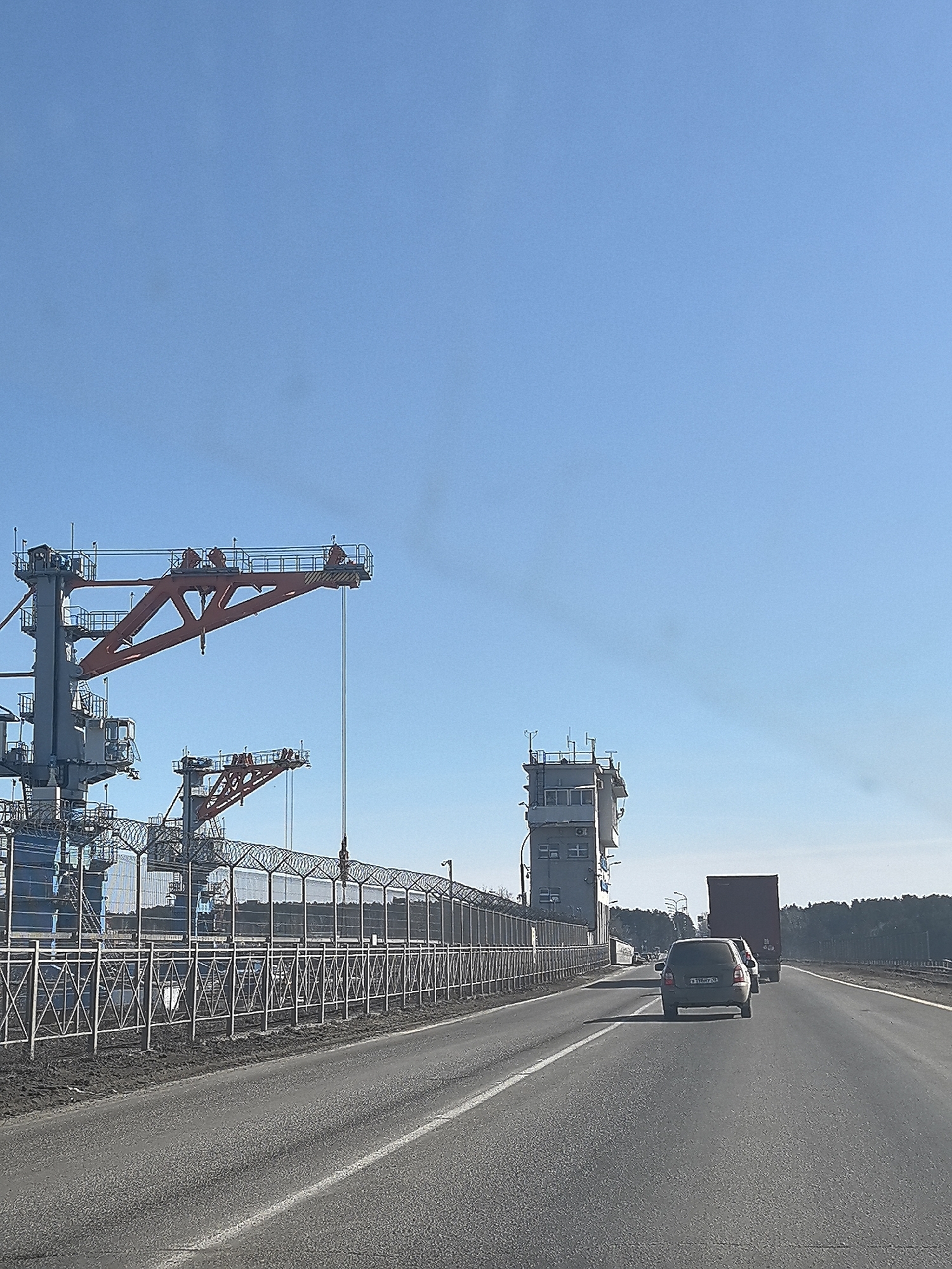 The image shows a highway scene in what appears to be the city of Chaikovsky, Russia. In the foreground, a two-lane road is visible, with a gray car and a red truck traveling away from the viewer in the right lane. To the left of the road is a tall, elaborate metal fence topped with barbed wire. Behind the fence is a large industrial structure consisting of cranes, towers, and buildings. The sky is clear and blue. There is a small amount of smoke emanating from behind the structure and to the left. The background features a treeline in the distance.