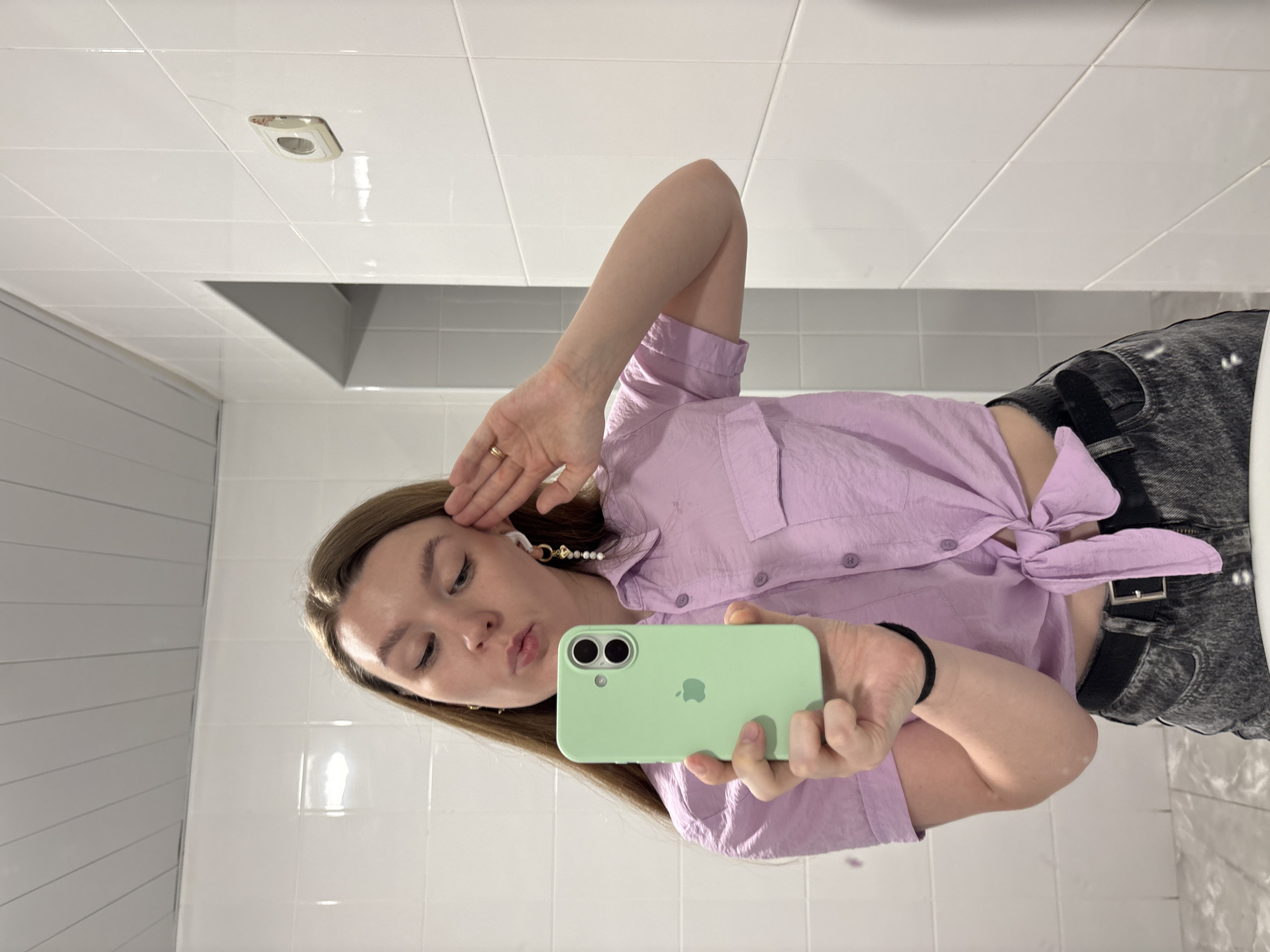 An indoor, mirror selfie captures a young woman posing playfully in what appears to be a bathroom or tiled room in Izhevsk, Russia. The woman, with long light brown hair, is looking directly at her phone screen, her lips slightly puckered. She is holding a light green iPhone, visibly featuring the Apple logo on its case, in her right hand. Her left hand is raised to her head, covering part of her ear and hair, and she wears a simple ring on her left ring finger. A decorative white wireless earphone, adorned with pearls and a gold accent, is visible in her left ear.
She is dressed in a light purple, collared, short-sleeved shirt, tied in a knot at the front to expose her midriff. She also wears dark grey or black acid-wash jeans or shorts with a black belt. A black hair tie is on her right wrist. The background consists of white rectangular tiles covering the walls and ceiling. A dark, recessed area, possibly a shelf or part of a vanity, is visible in the background, along with a rectangular ventilation fan or light fixture on the ceiling. The lighting suggests an artificial indoor environment. The image itself is rotated 90 degrees counter-clockwise.