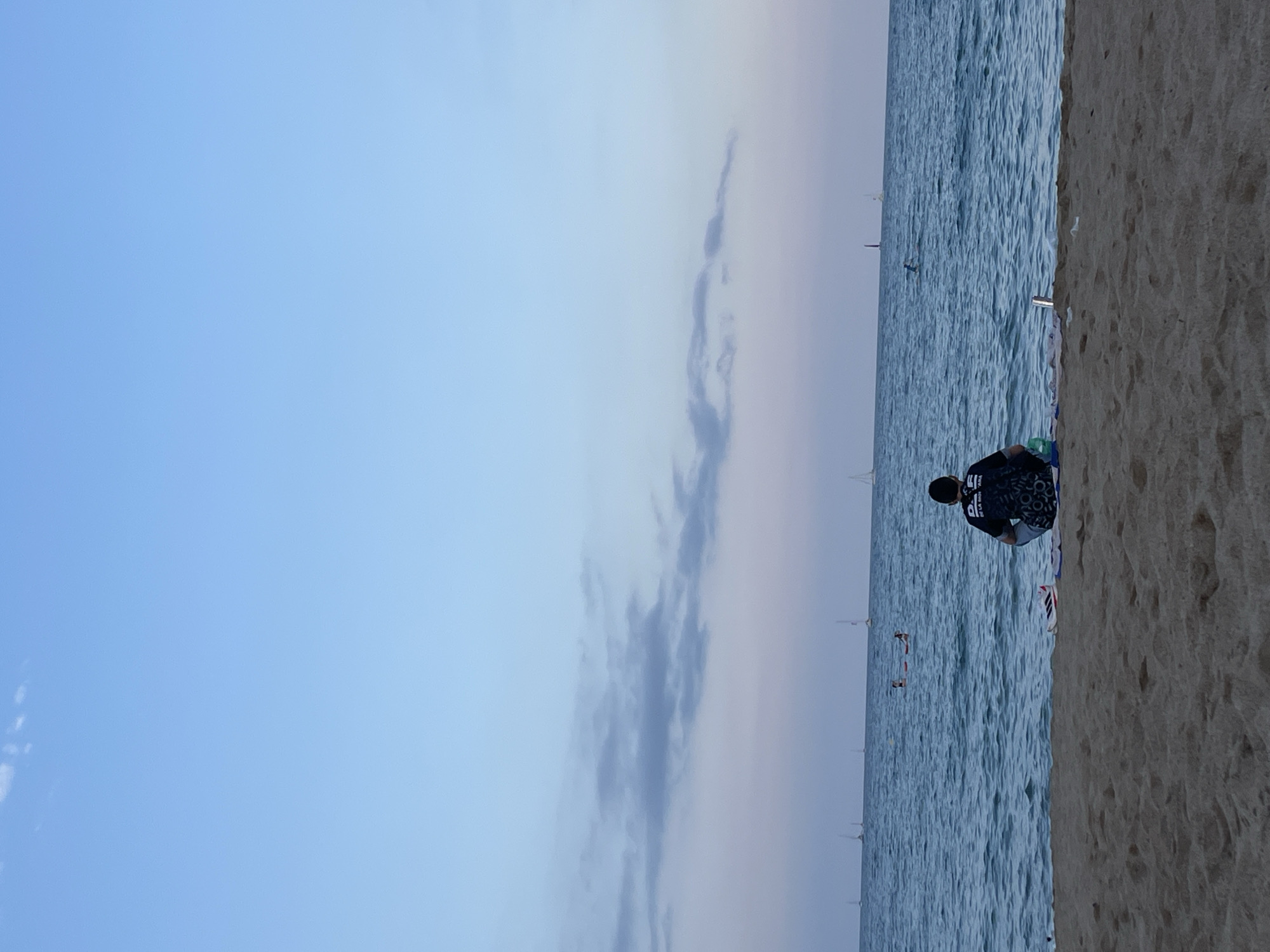 A person sits facing the ocean on a sandy beach. The person is wearing a dark t-shirt and a dark cap. The person is sitting with their legs crossed, looking out at the water. In the distance, there are several sailboats on the ocean. The sky is a gradient of light blue to a pale pinkish hue, with some scattered clouds. The beach sand is light brown and textured. There are a few items on the sand near the person, including a pair of shoes and a blue bag. This image was taken at Barceloneta Beach in Barcelona, Spain.