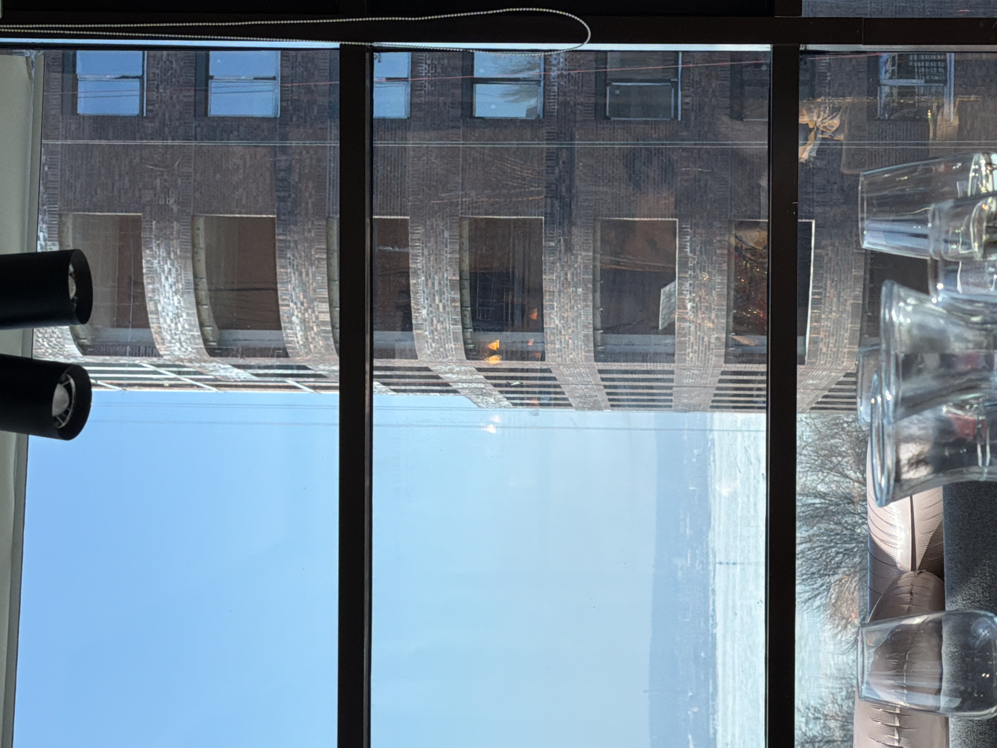 The image presents an elevated interior view looking through a multi-paned window onto an urban and natural landscape. The location is Izhevsk, Russia, specifically overlooking the Izhevsk Pond, with a prominent view of a tall, curved brick building identifiable as the "Atlant" residential complex at Kirova Street 8A.
In the immediate foreground, a partial indoor scene is visible. Several clear glass vessels, including at least two wine glasses and multiple tumblers, are arranged on a dark, textured surface. Behind these, a segment of a light-brown, horizontally-ribbed, padded banquette or backrest is partially shown. Two dark, cylindrical overhead light fixtures are suspended from the ceiling in the upper portion of the frame. Dark horizontal and vertical window mullions divide the view, and a white beaded chain hangs vertically along the right-hand window frame.
The midground is dominated by a multi-story building constructed of dark-red brick, featuring a distinctive curved facade with recessed, arched, or rectangular openings across its levels. Some of these openings contain installed windows with light-blue tinted glass, while others remain open, revealing interior construction and structural elements. Reflections of interior light sources or parts of the room are visible on the window glass.
The background reveals a wide panorama under a clear, bright blue sky. A large, frozen body of water, the Izhevsk Pond, stretches horizontally, appearing white and textured. Beyond the frozen surface, a distant landmass with a hazy outline of bare trees and faint structures is discernible along the horizon. No individuals are visible in the scene.