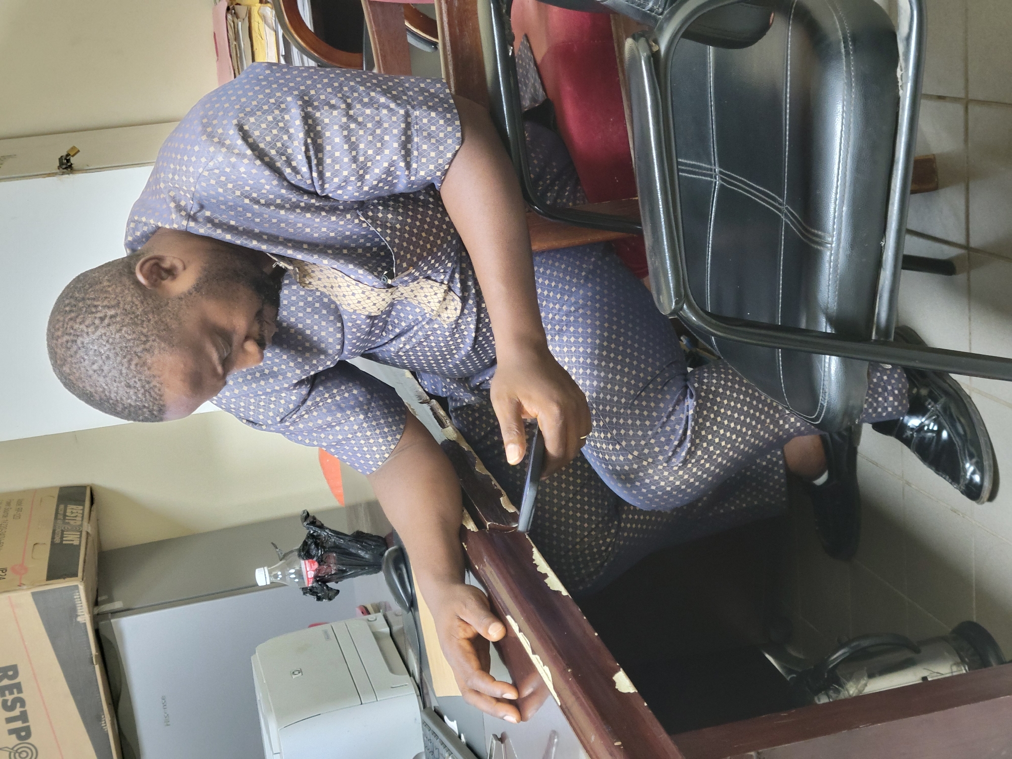 An adult male is seated, focused on meticulously removing paint from a wooden object with a sharp tool. The man is wearing a patterned, light blue and gold ensemble. He is seated on a chair, next to a padded chair with a metal frame and black leather. He is wearing black leather shoes and white socks.
To the left of the man, there is a desk with a printer, water bottle, and trash bag. Above the desk there is a cardboard box labeled "RESTPOINTE". The flooring and lighting indicate an interior space. There is no information to confirm this image was taken in the city of Abuja, Nigeria.