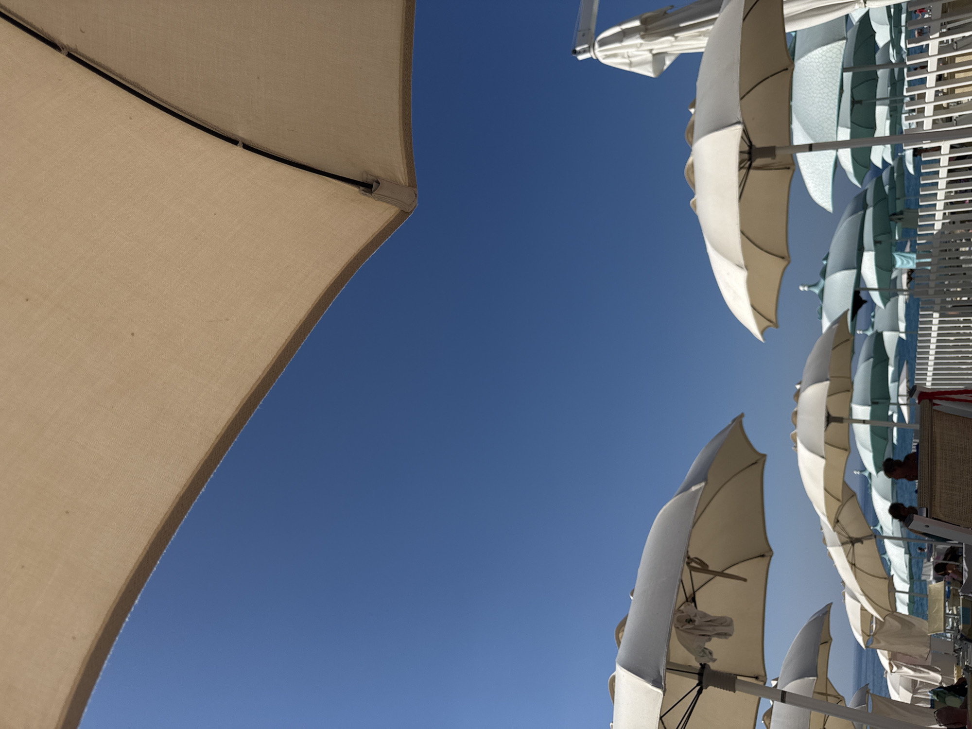 This image depicts a beach scene in Finale Ligure, Italy, dominated by a clear blue sky and numerous beach umbrellas. In the foreground, a large, tan umbrella occupies the upper portion of the frame, casting a shadow against the sky. Below this, a row of white and cream-colored umbrellas are clustered together, some with their fabric coverings partially visible. Further back, a variety of other umbrellas, including some in light blue and white, are spread across the beach. The sea is visible in the background, appearing calm and a deeper blue than the sky. Several people are present on the beach, some seated under the umbrellas or walking amongst them. A white picket fence runs across the lower part of the image, separating the seating area from the sand and sea. The overall impression is of a sunny, leisure-filled day at the beach.
