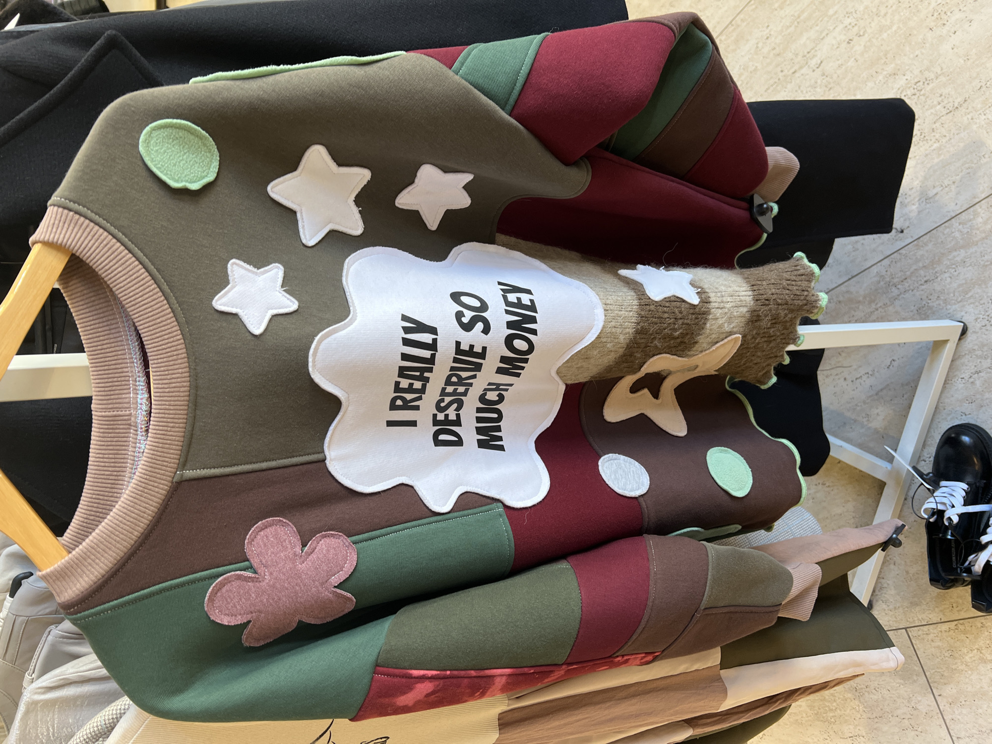 The image depicts a colorful, patchwork-style sweater with appliqued shapes and text, resting on a wooden hanger amidst a collection of clothing on what appears to be a rack or display. The sweater features a light green circle, several white star shapes, and a white cloud-shaped patch with the text "I REALLY DESERVE SO MUCH MONEY" printed in black. The color scheme is a mix of earth tones like olive green and brown, interspersed with sections of burgundy and lighter greens. A pink flower applique is located in the lower left corner of the garment. The garment is made of various fabric patterns and textures including a brown, tan and white striped sleeve. Part of a black jacket and other clothing items are visible in the background. A pair of black shoes, likely belonging to a mannequin or customer, are on the right of the frame. The overall impression is a retail setting, suggesting a clothing store or boutique. I cannot verify the location.