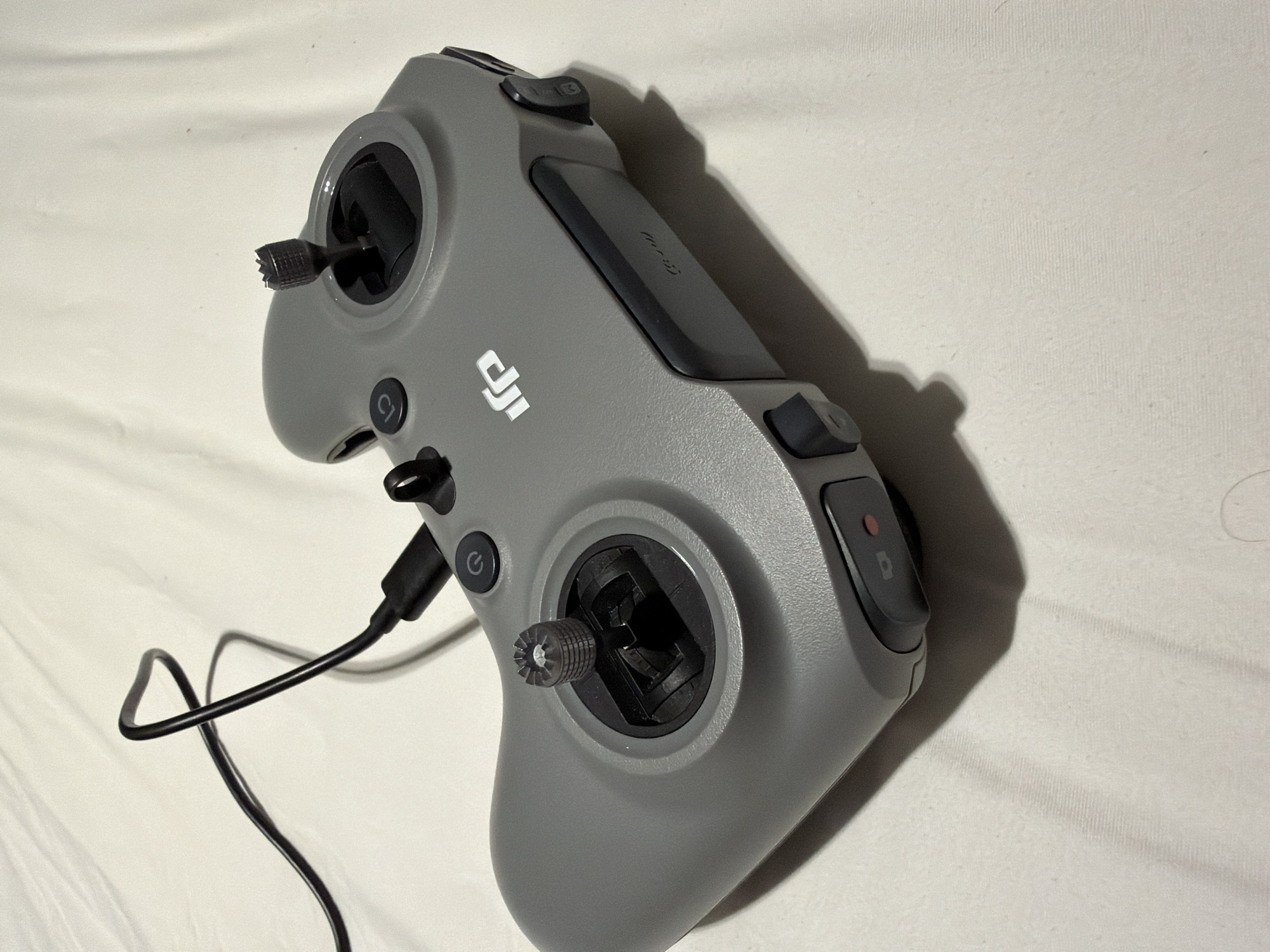 The image displays a gray DJI remote controller on a white, textured surface, likely a bedspread. The controller features two joysticks, several buttons, and a USB-C port with a black cable connected. The DJI logo is visible on the front of the controller. The lighting suggests an indoor setting, with a shadow cast by the controller. No people are present in the image. The location in Ban Phonsavang Neua, Laos, is not discernible from the visual information provided in the image.
